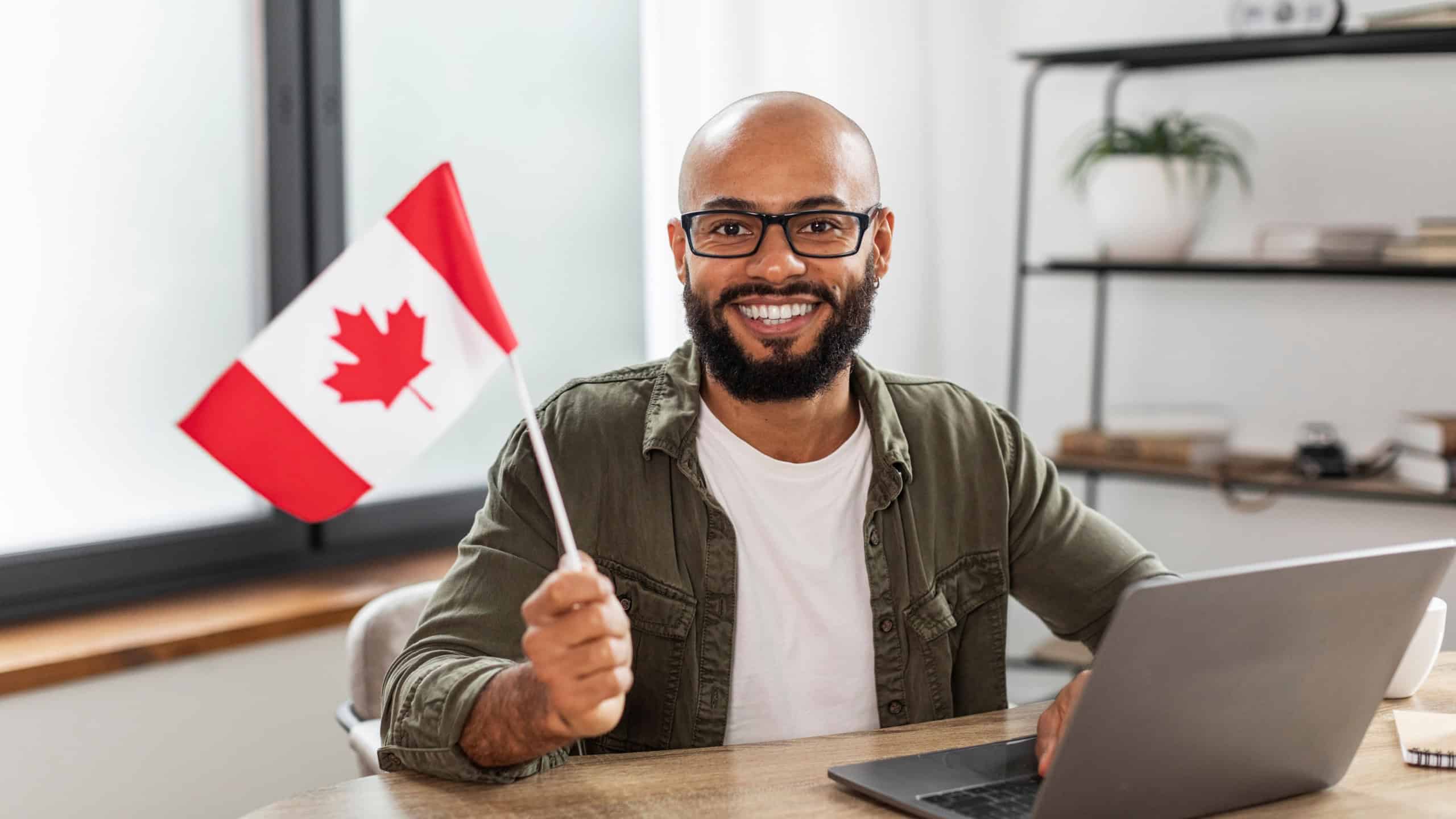 Happy latin man sitting with flag of Canada and using laptop computer, sitting at desk at home. Modern online foreign education, emigration and citizenship, working abroad concept