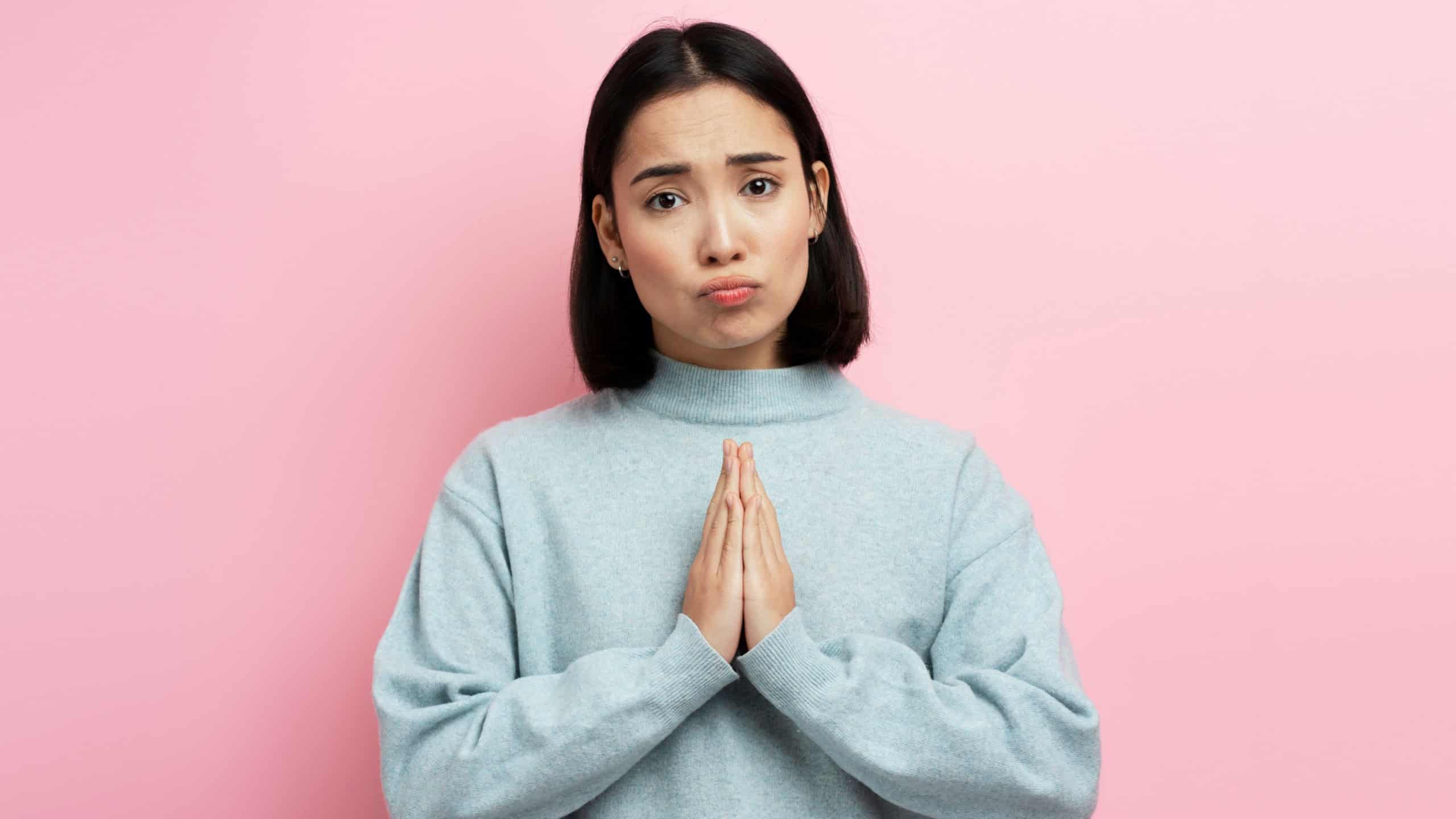 Portrait of attractive woman praying, begging with pleading grimace, gesture of asking apologising. Indoor studio shot isolated on pink background