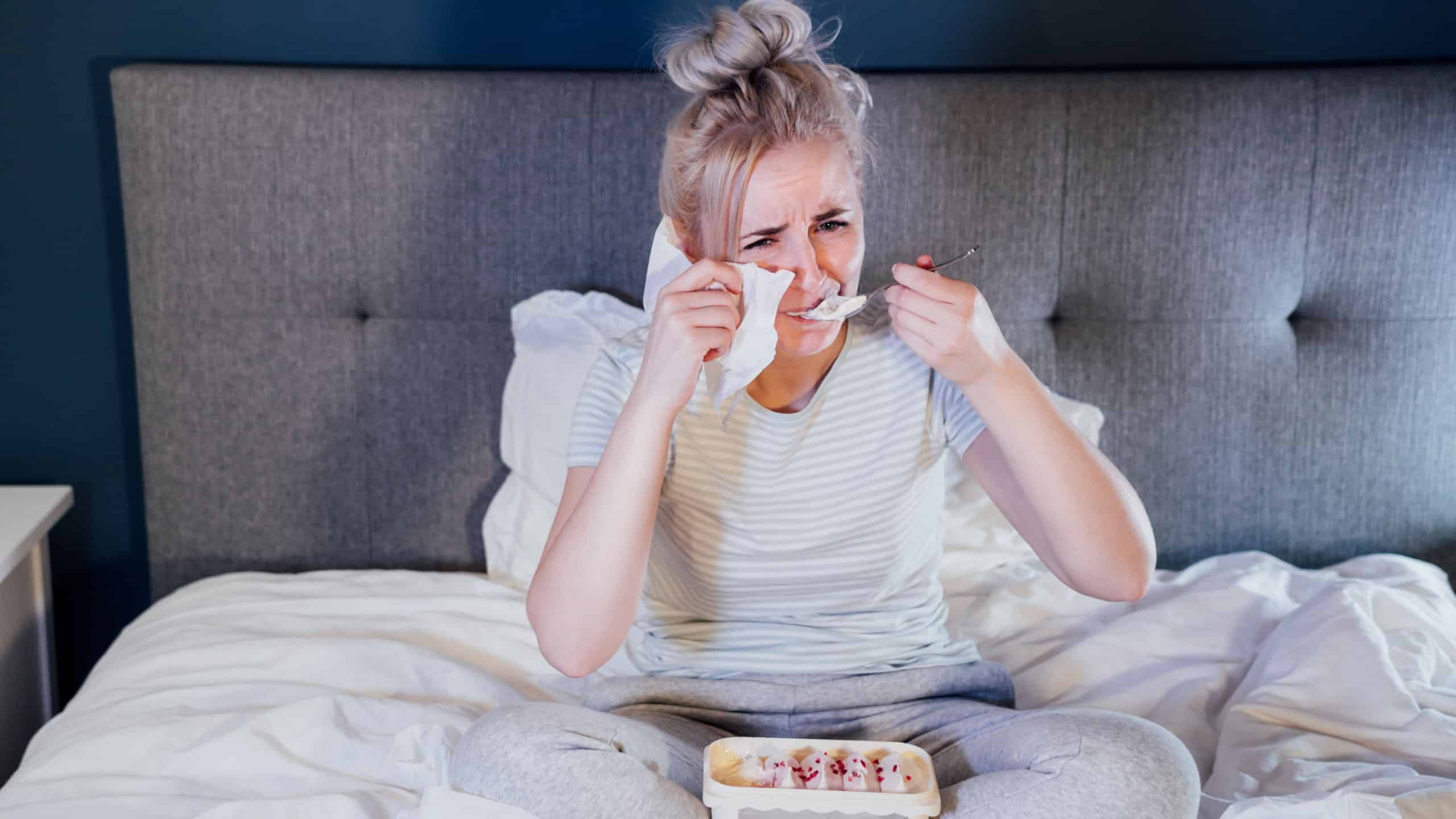 Upset young woman holding tissue, eating ice cream from bucket while watching romantic movie on TV at home. Sad lady crying over breakup or relationship problems, feeling depressed and lonely