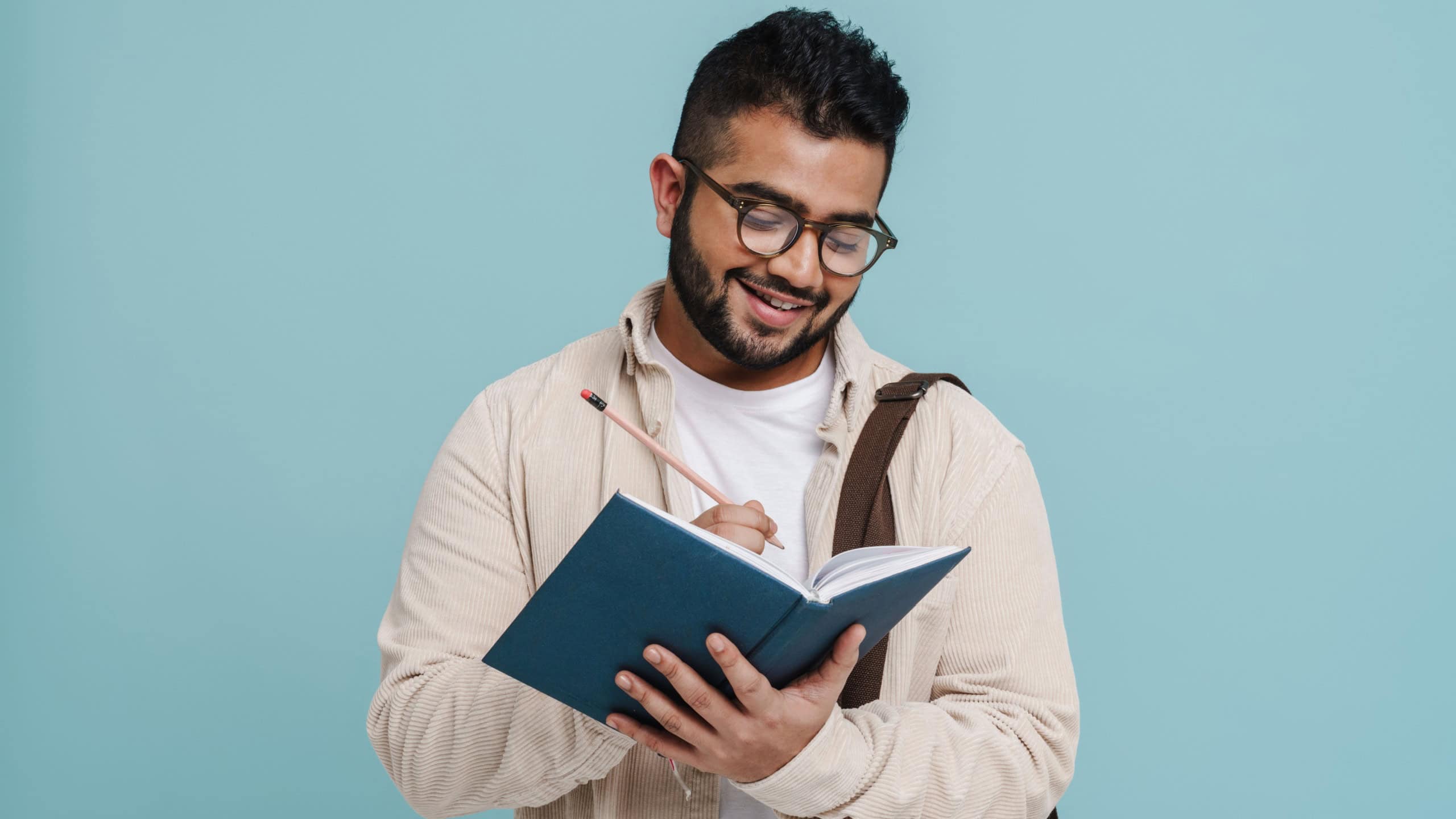 Smiling young indian man wearing eyeglasses writing down notes while standing isolated over blue studio background