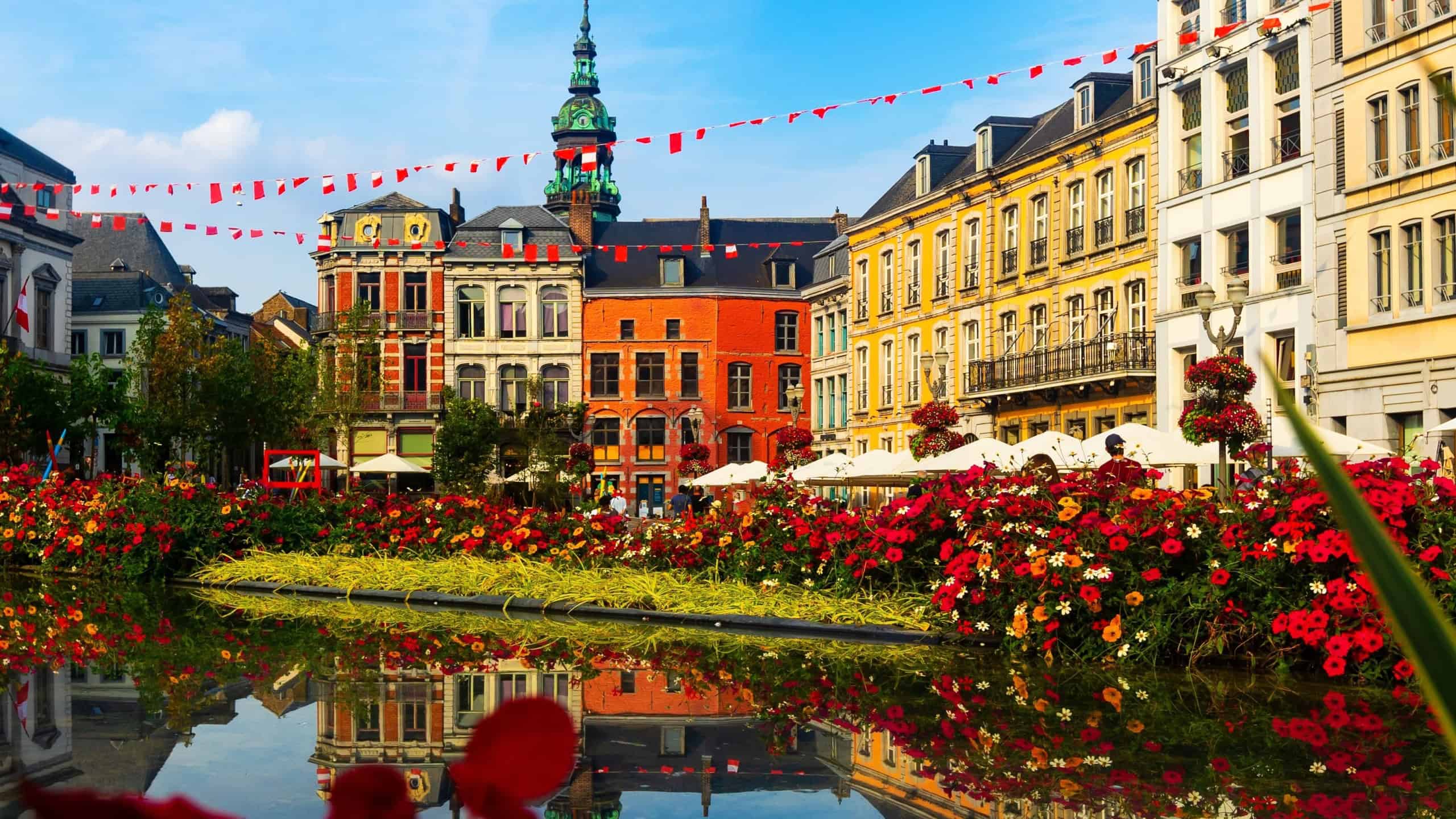 Picturesque summer view of flowering central Grand Place square in Mons overlooking baroque belfry of Roman Catholic Church of St. Elizabeth towering over colorful residential townhouses, Belgium