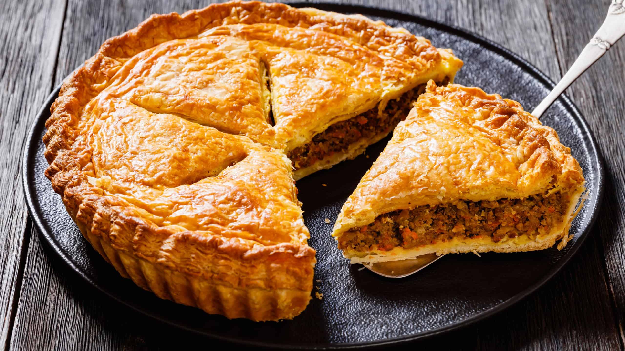 sliced Ground Beef Meat Pie with a flaky puff pastry double crust with hearty minced beef cooked with vegetables and seasoning on black plate on wood table, close-up