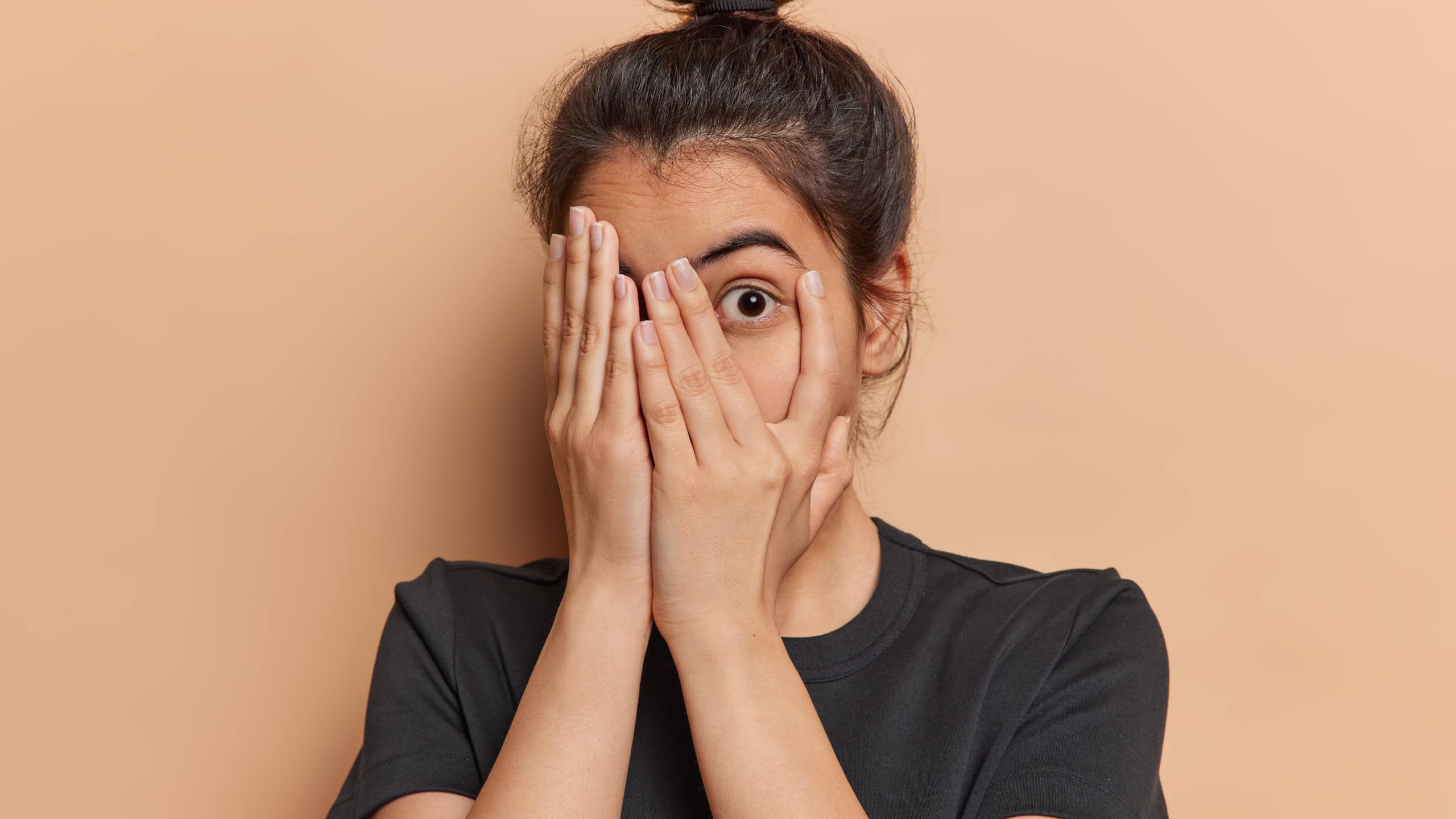 Scared Latin woman with hair bun peeks through her fingers fearfully hiding her face unable to bear sight of something terrible witnessing something truly terrible dressed in casual t shirt isolated