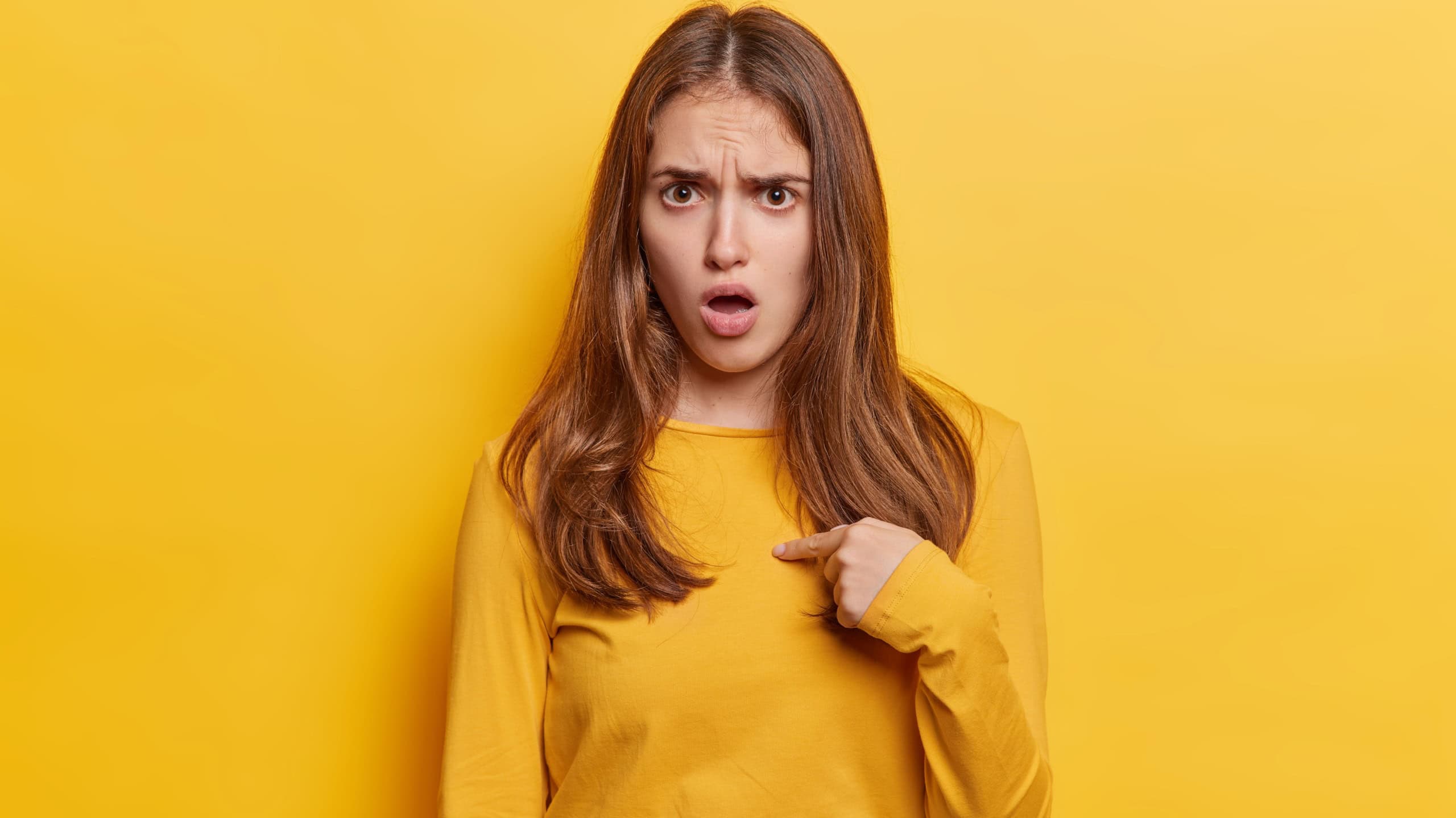 Young woman points finger at chest verbally defending herself with perplexed and puzzled expression with a challenging gaze and indignant tone exclaims who me dressed casually isolated on yellow wall