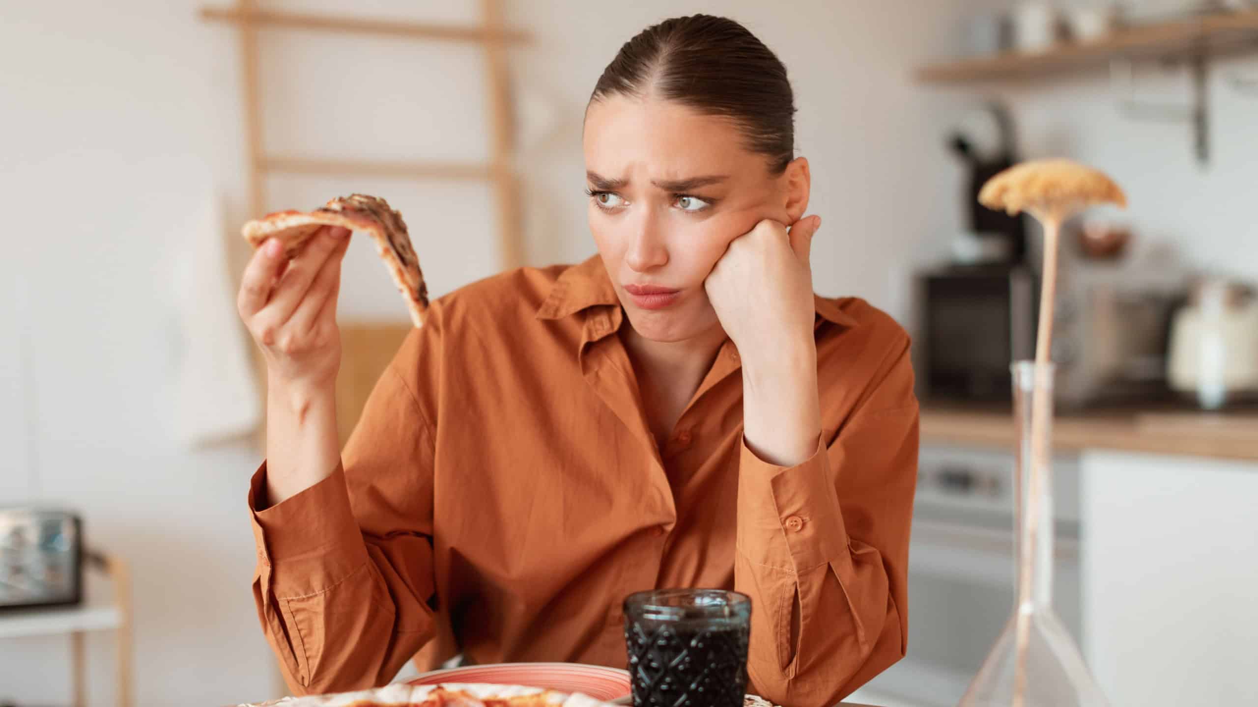 Upset woman looking at delicious piece of high-calorie pizza, holding slice in hand. Diet and digestive problems as well as spoiled and tasteless food