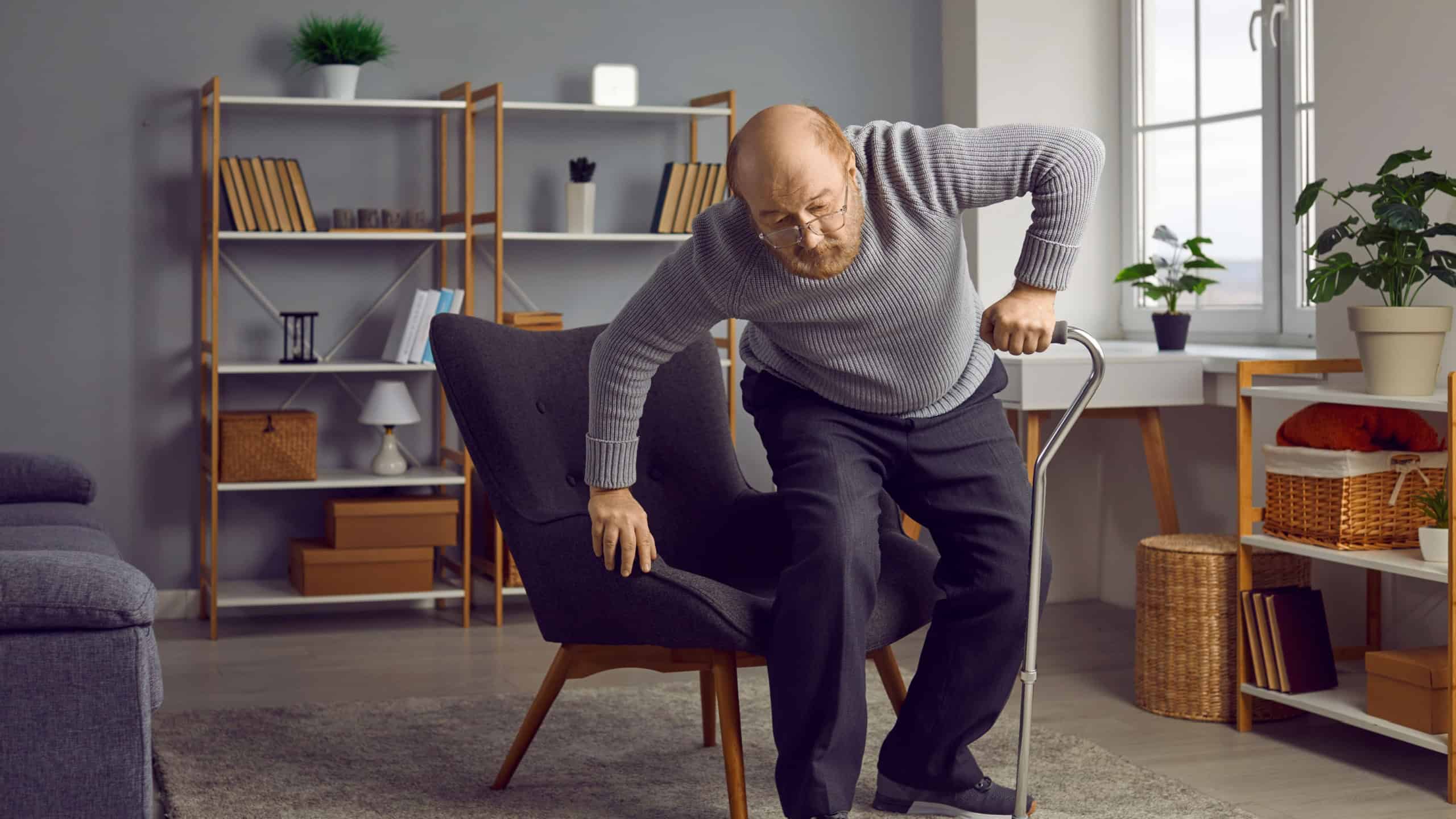 Retired old man with aching joints uses a cane to walk. Bald senior man with a walking stick stands up from an armchair in the living room at home