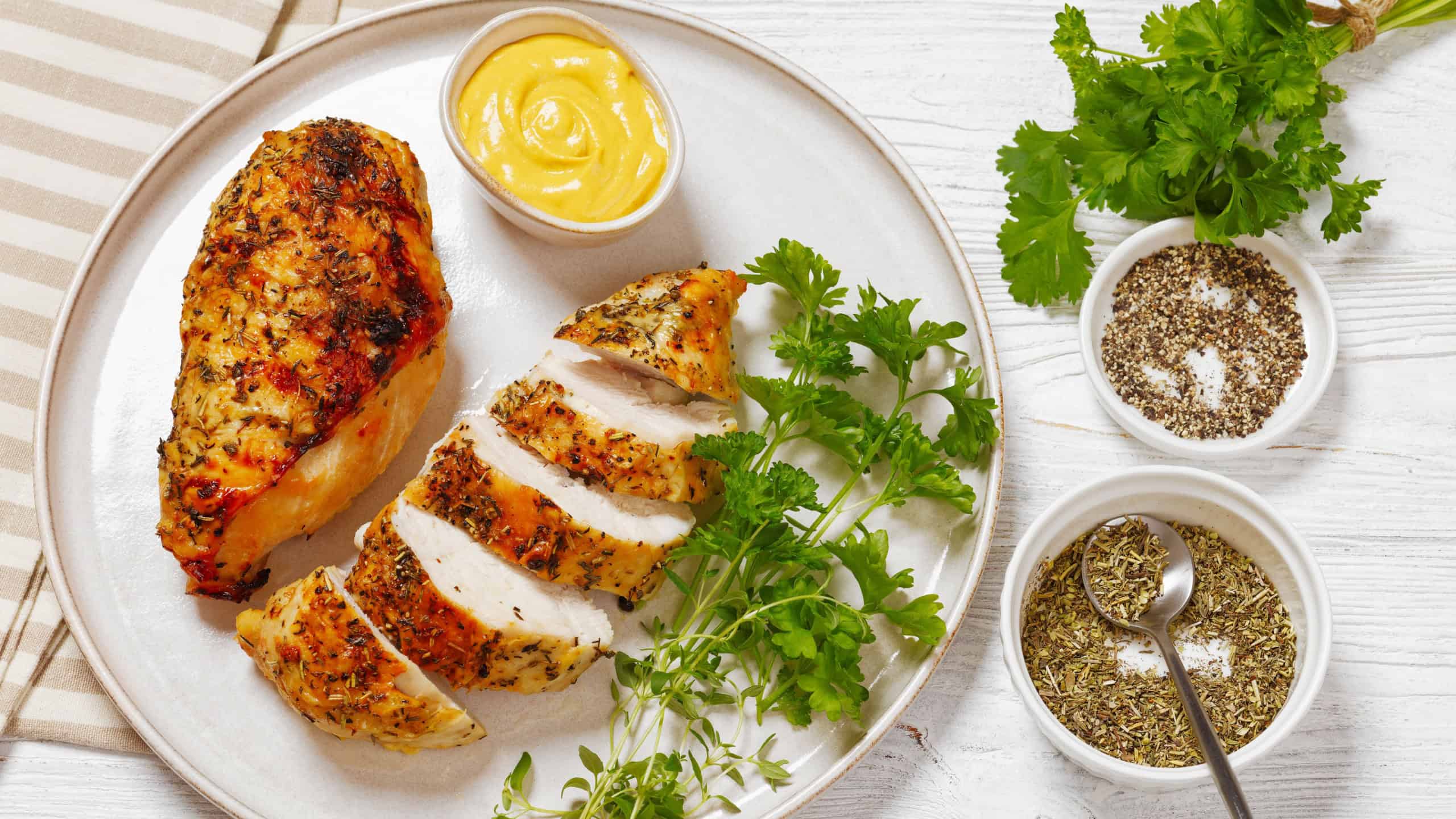 Baked Split Chicken Breasts with mustard, parsley and thyme on plate on white wooden table with cutlery and seasoning
