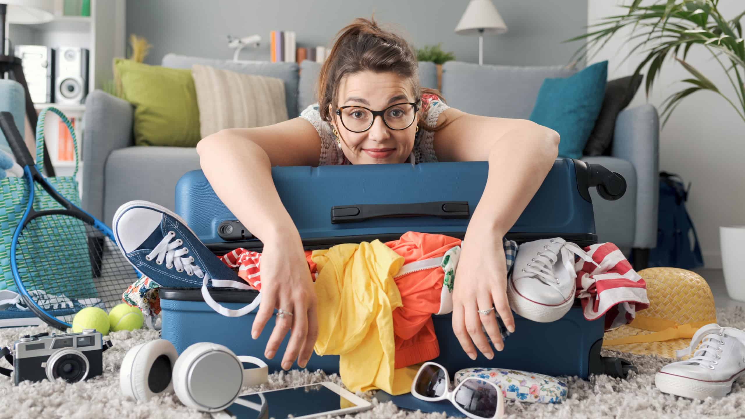 Happy young woman packing for a trip, she is leaning on her full trolley suitcase and smiling