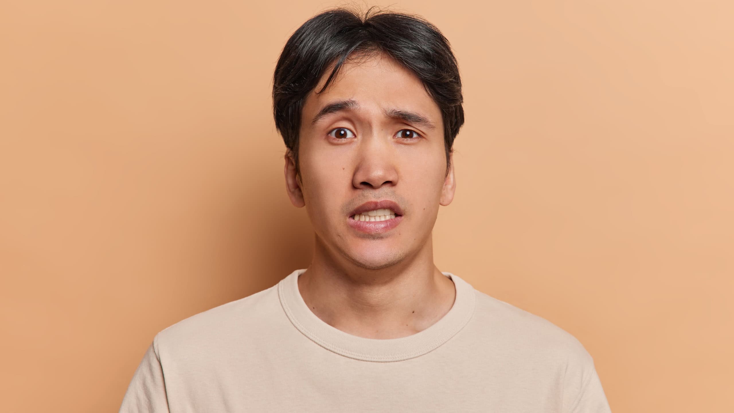 Human facial expressions concept. Brunet young Japanese man clenches teeth and looks embarrassed at camera dressed in casual t shirt dressed in casual t shirt isolated over brown background.