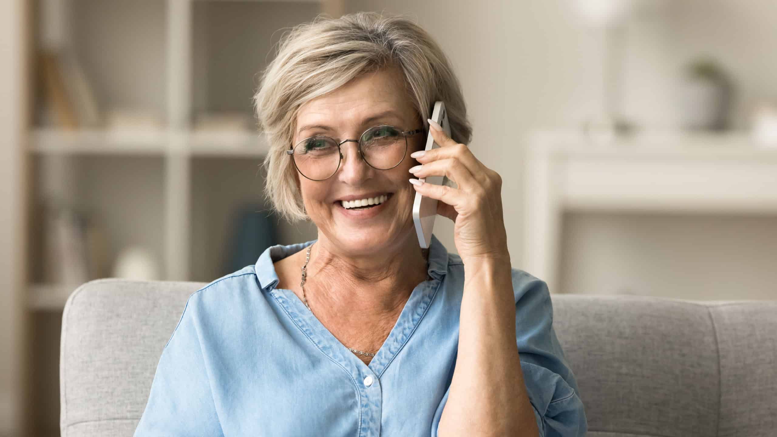 Cheerful pretty blonde senior woman in stylish eyeglasses enjoying telephone call, connection, funny conversation on home sofa, looking away with toothy smile, listening, speaking, laughing
