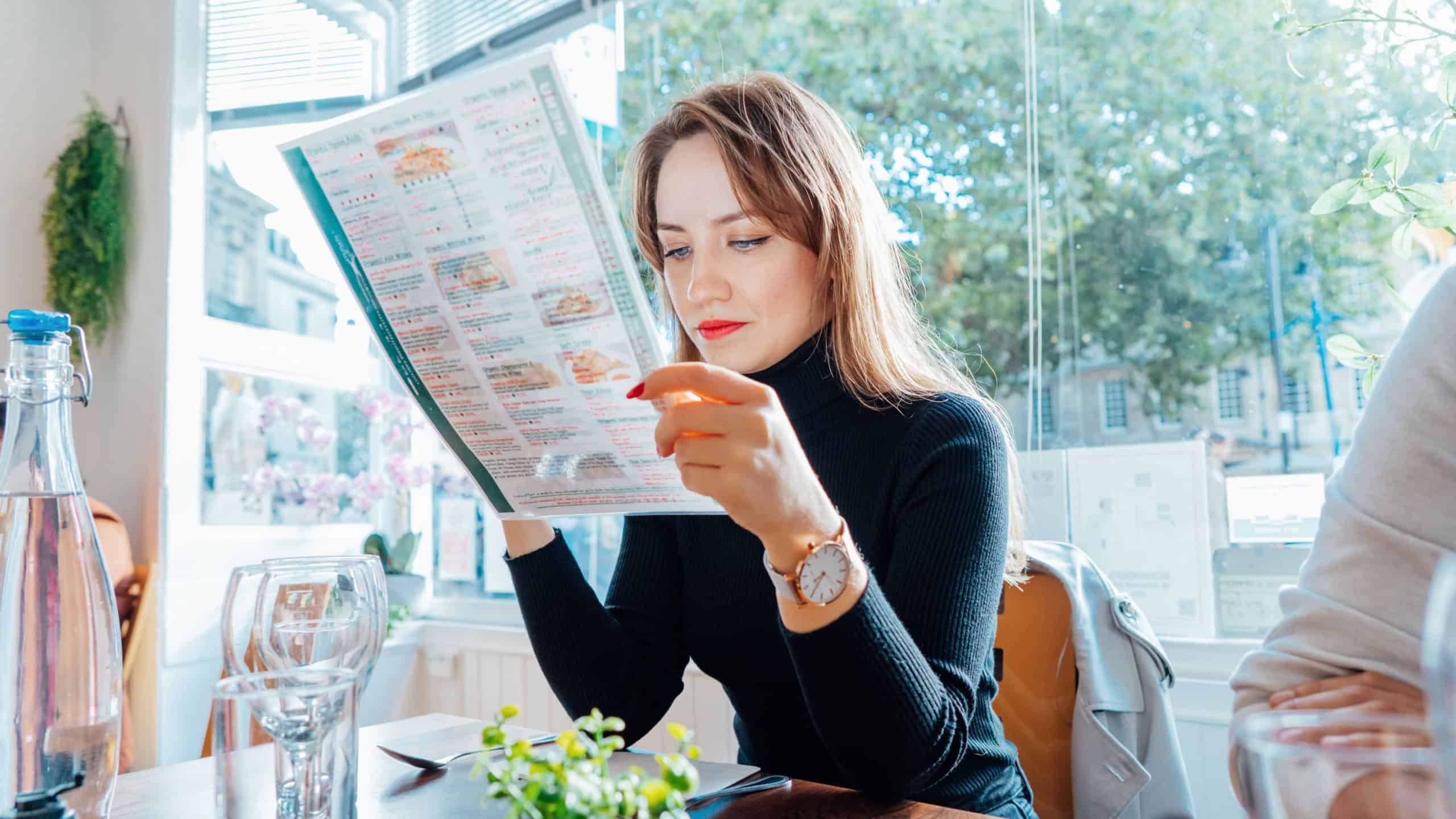 Young woman reading menu sitting with friend at casual cafe. Smiling woman checking menu, choosing meal or drink, making order. Try new foods while traveling. real people. copy space. Selective focus.