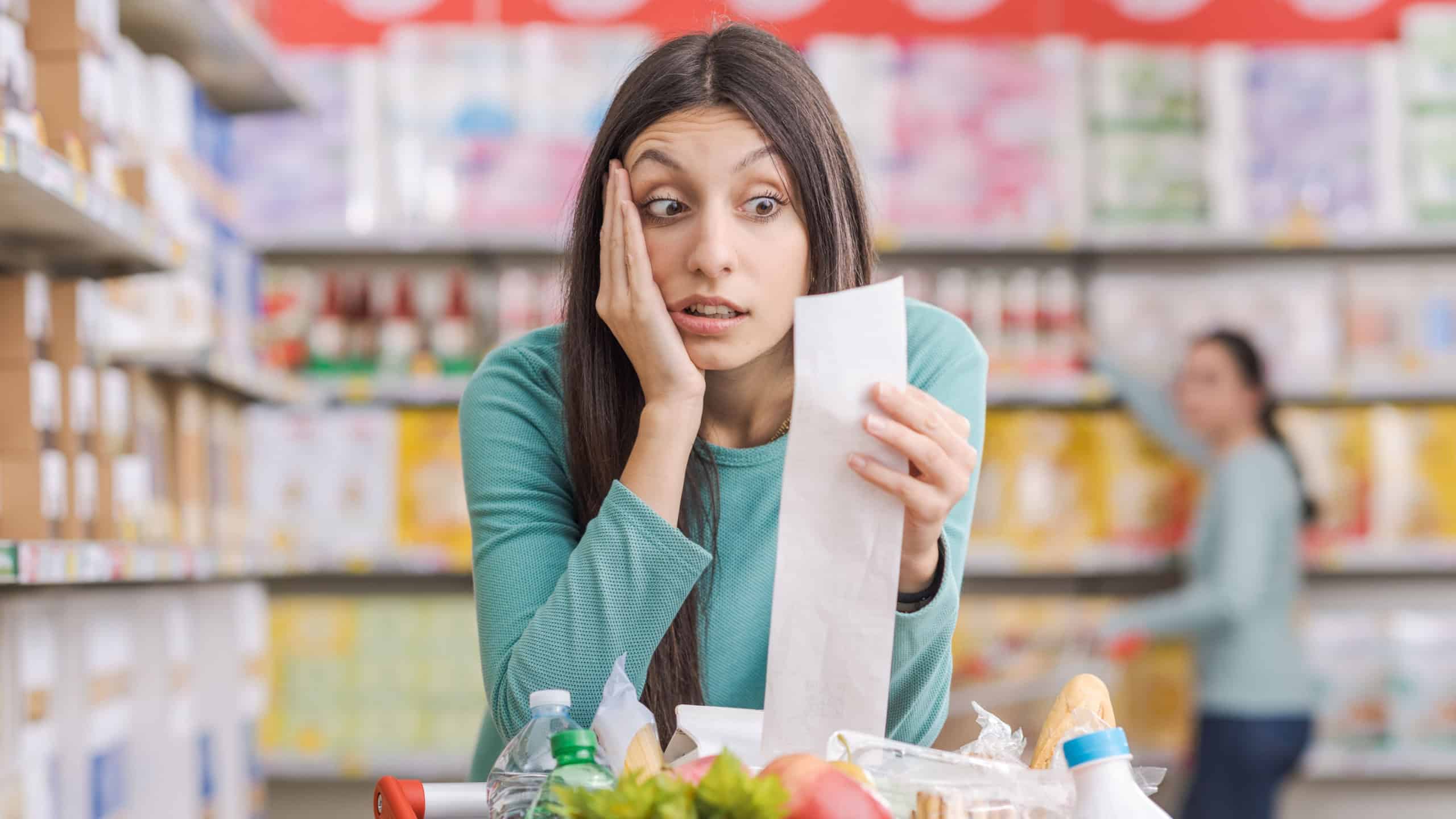 Young shocked woman at the supermarket, she is leaning on a shopping cart and checking a long expensive grocery receipt