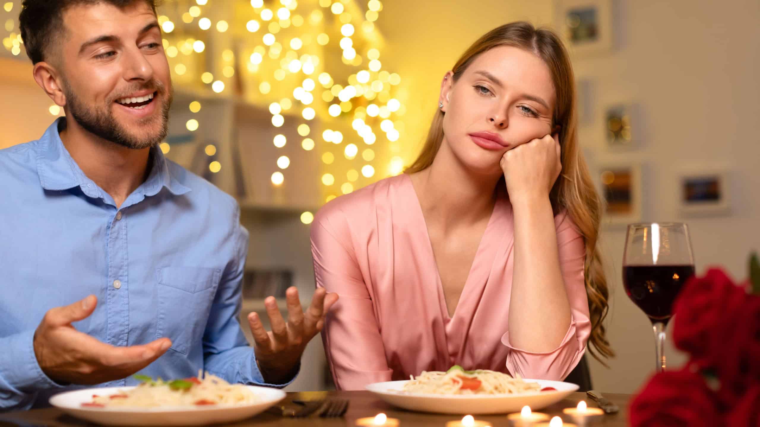Disinterested woman rests her cheek on her hand at dinner table, looking away from excited man speaking passionately, highlighting stark contrast in their moods