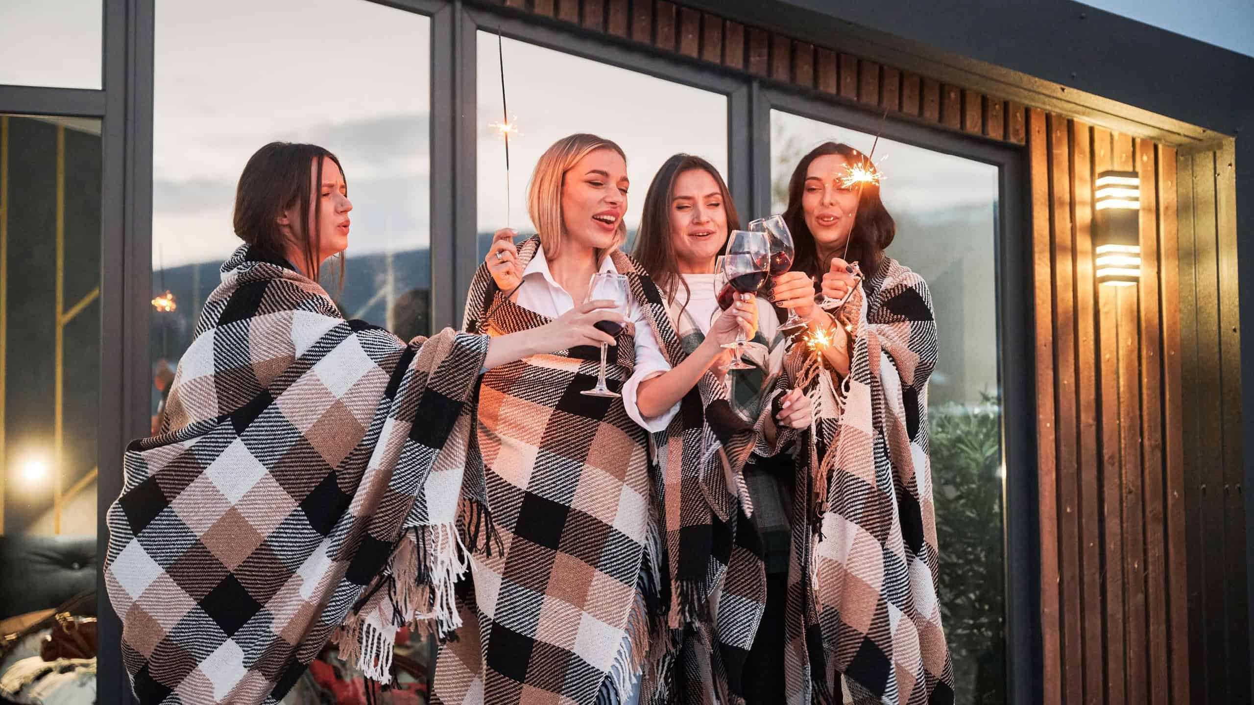 Young women enjoying winter weekends on terrace of contemporary barnhouse. Four girls in plaids drinking wine and celebrating with sparklers in the evening.