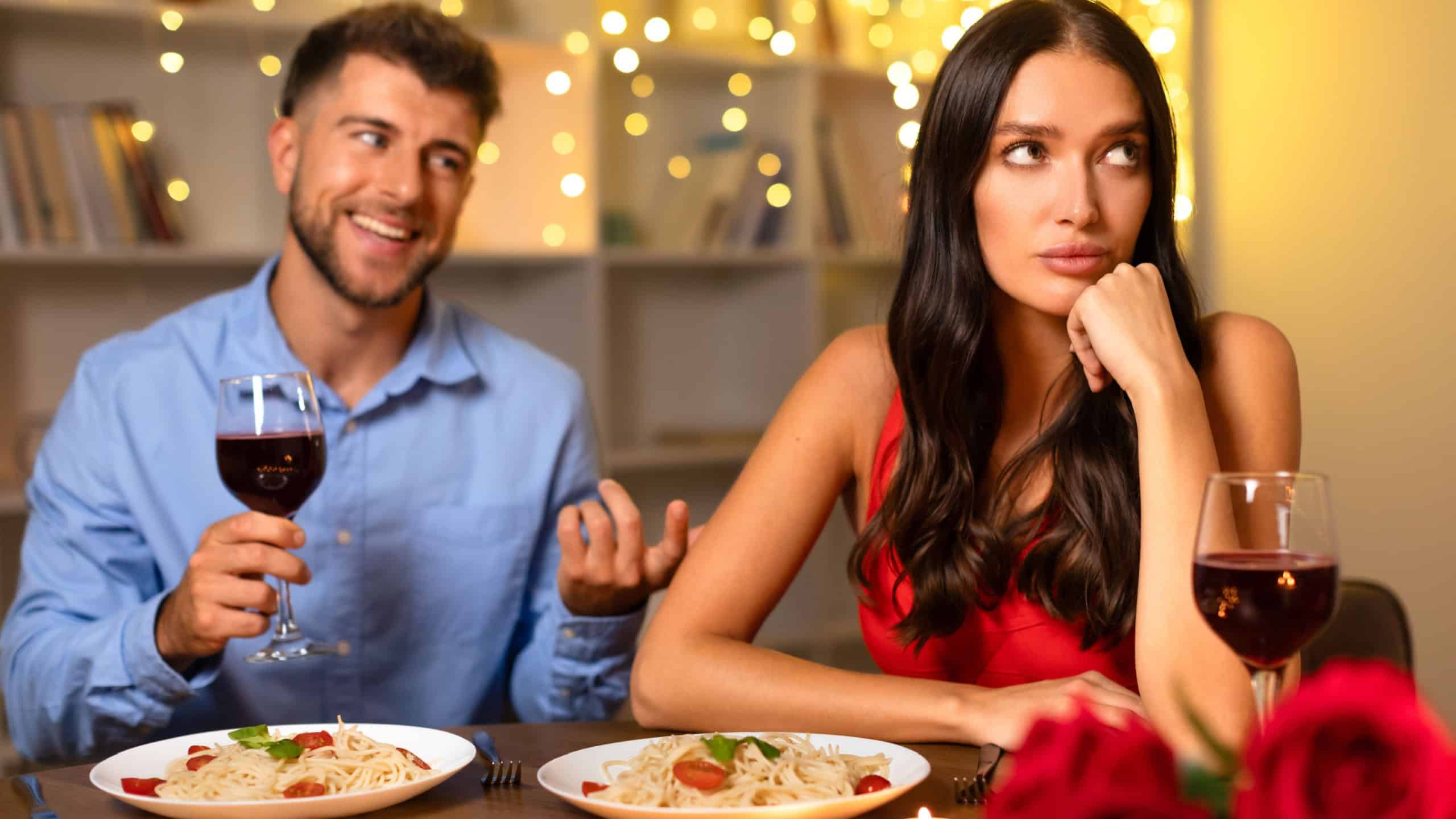 Cheerful man talking and holding glass of wine, while the woman beside him looks away in disinterest, amidst romantic dinner setting