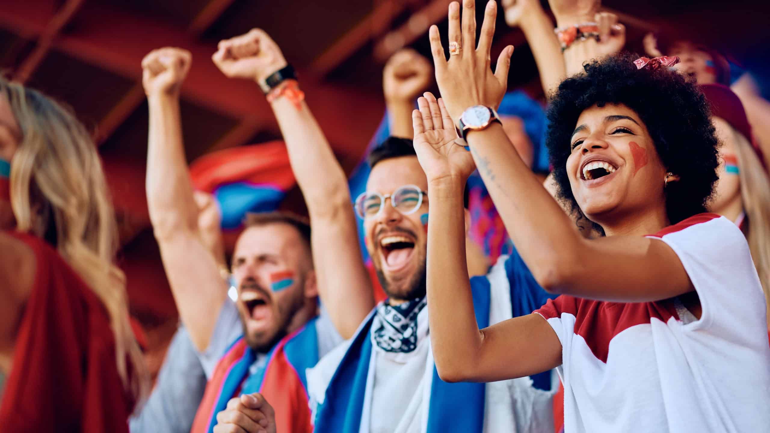 Group of cheerful sports fans celebrating victory of their favorite team while spectating game from stadium stands. Focus is on African American woman.