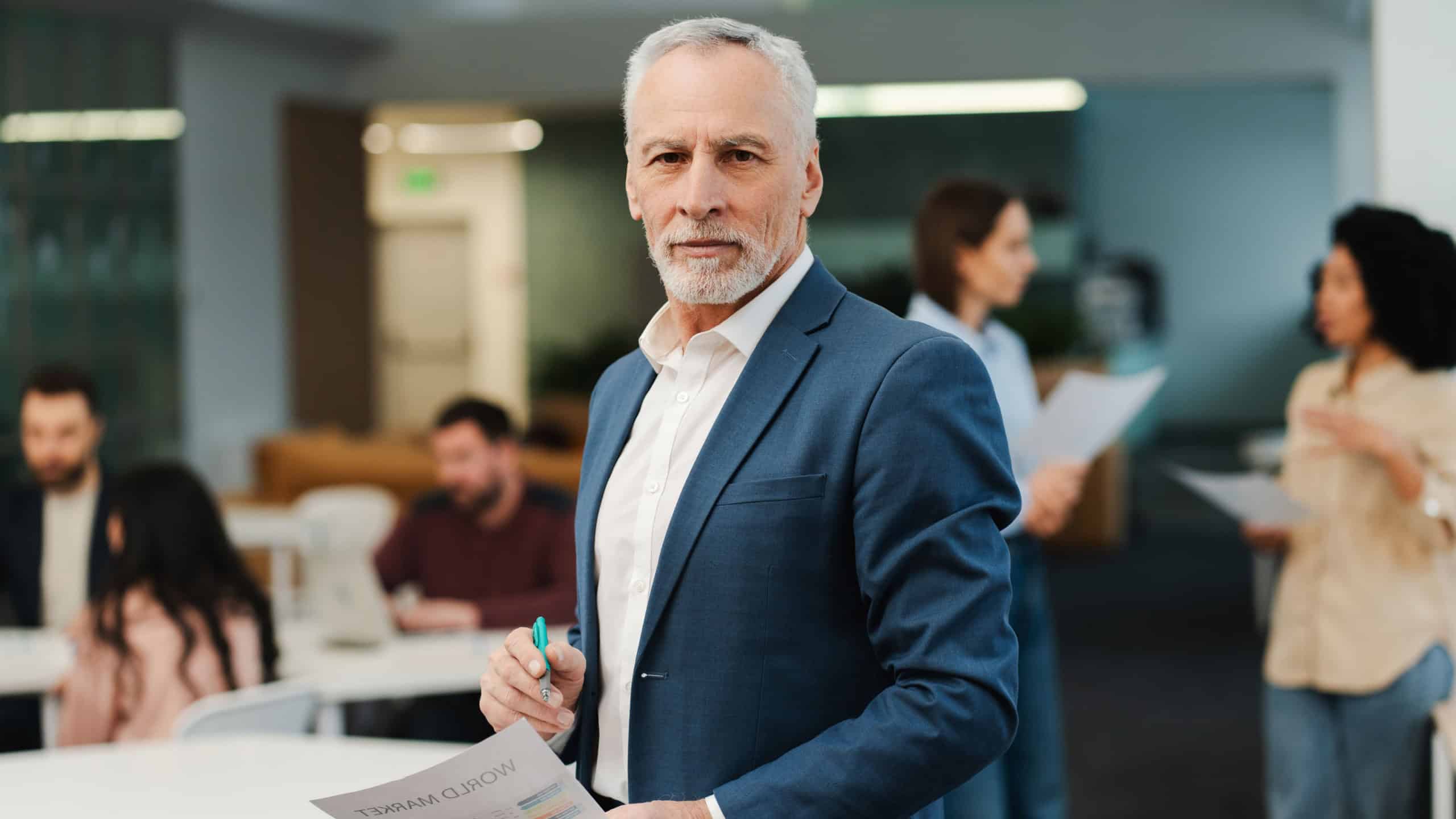 Attractive serious senior man, mature businessman wearing stylish suit looking at camera working with colleagues in modern office. Concept of successful business