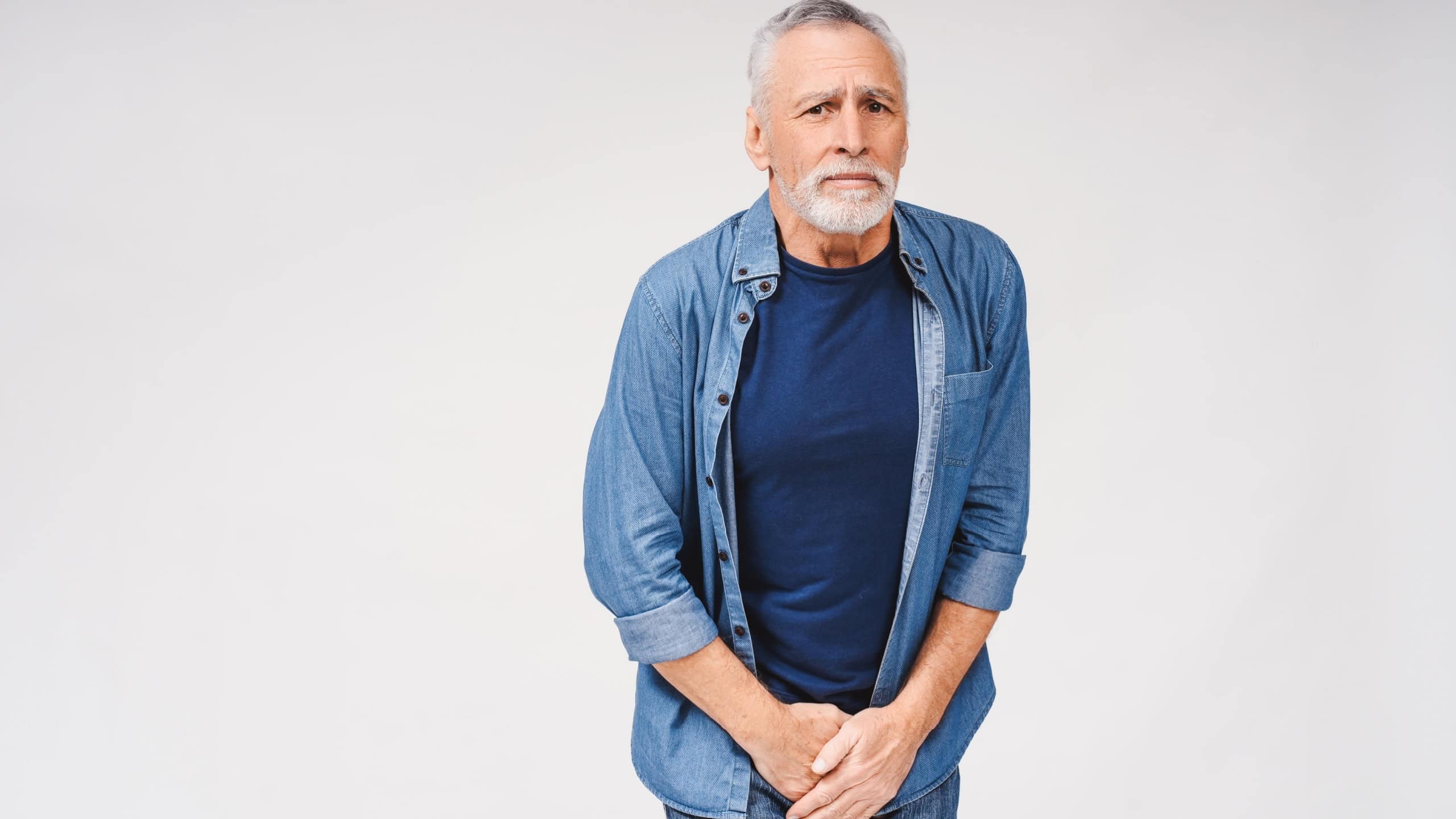 Portrait of senior gray-haired man holding hands at the genitals while posing at the studio with white background. Urological diseases, pain during urination concept