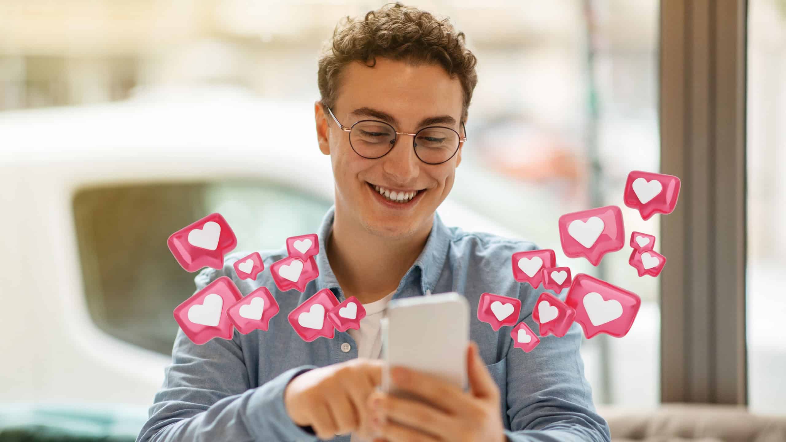 A man wearing glasses smiles broadly as he looks at his phone. A stream of social media heart icons, in a white heart inside a pink box, float around him. He is seated indoors, possibly in a cafe