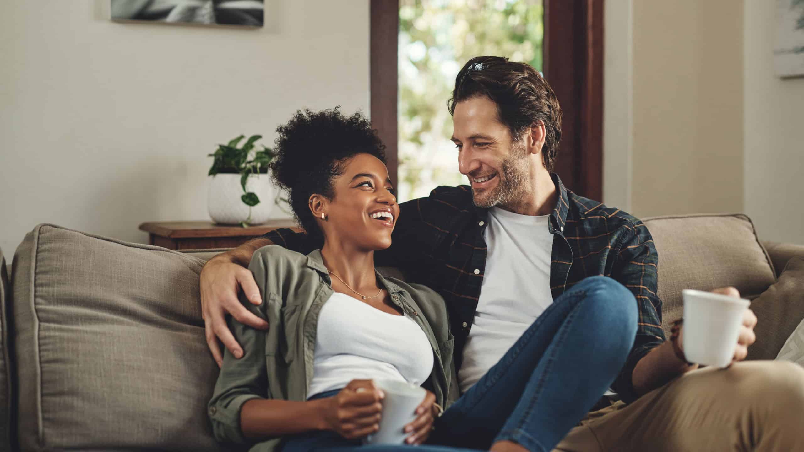 Happy, interracial couple and drinking coffee in home, talking or bonding together for love in living room. Tea cup, man and funny woman in conversation for joke, story or laughing at comedy on sofa
