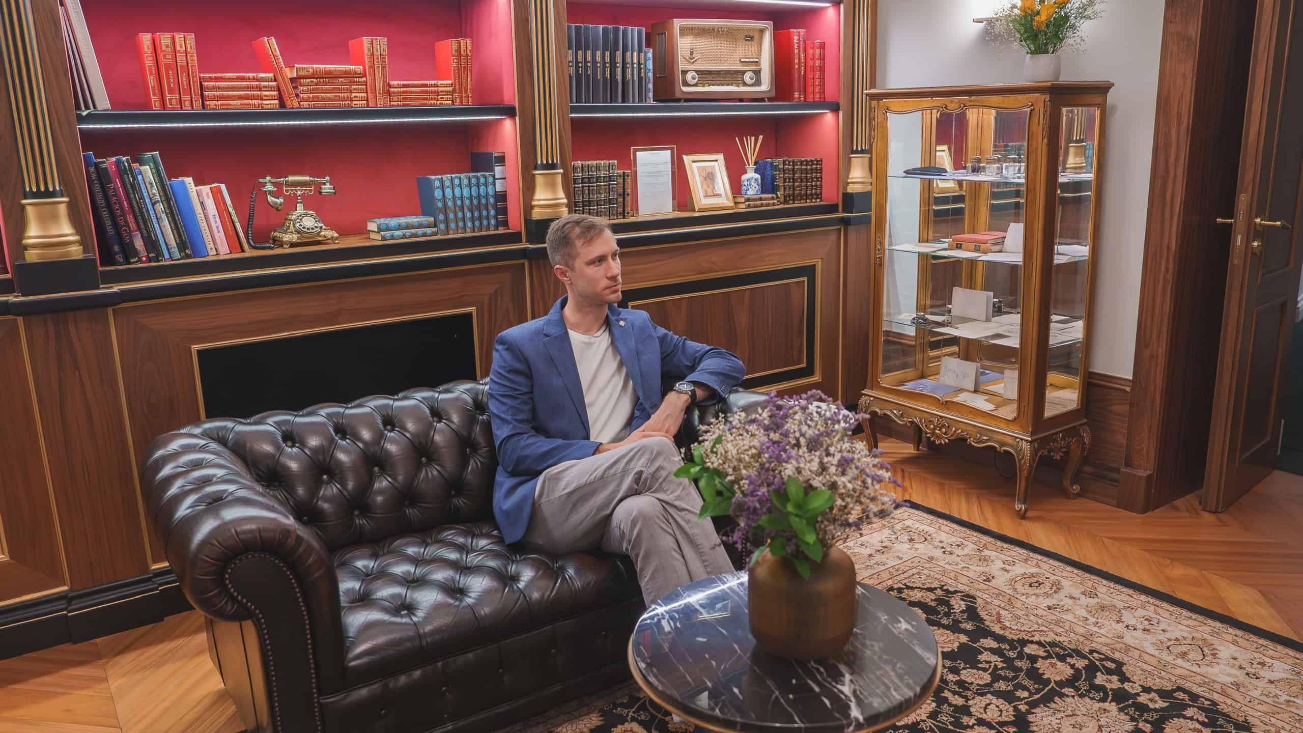 A man sits on a dark leather Chesterfield sofa in a luxurious hotel interior. The room features a red backlit bookshelf with vintage books, an antique radio