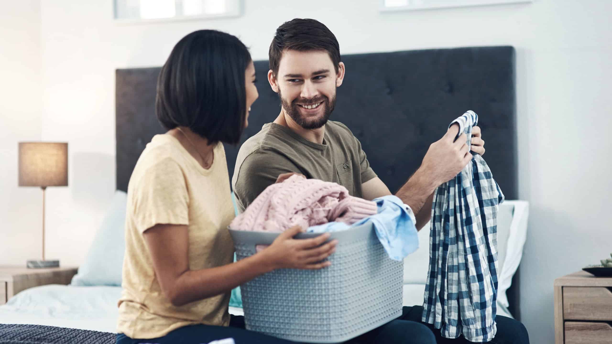 Couple, help and folding laundry on bed for support, shared chores and responsibility in home. Happy man, woman and clothes in bedroom for spring cleaning, sorting and routine housework on weekend