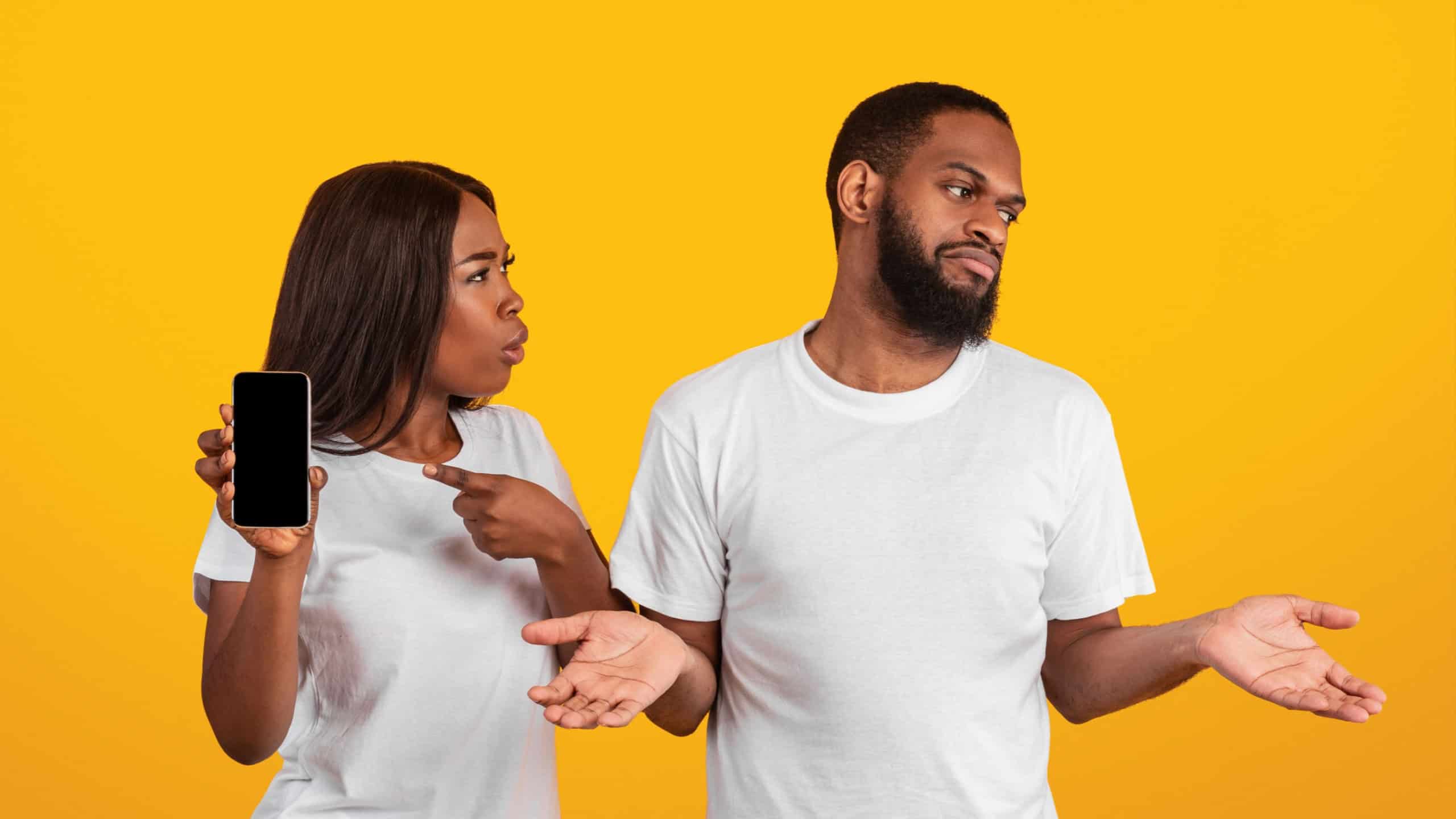 Gadget and Social Media Addiction, Cheating Concept. Angry black woman checking her boyfriend mobile phone, showing provocative messages and asking man who spreading hands, yellow studio background
