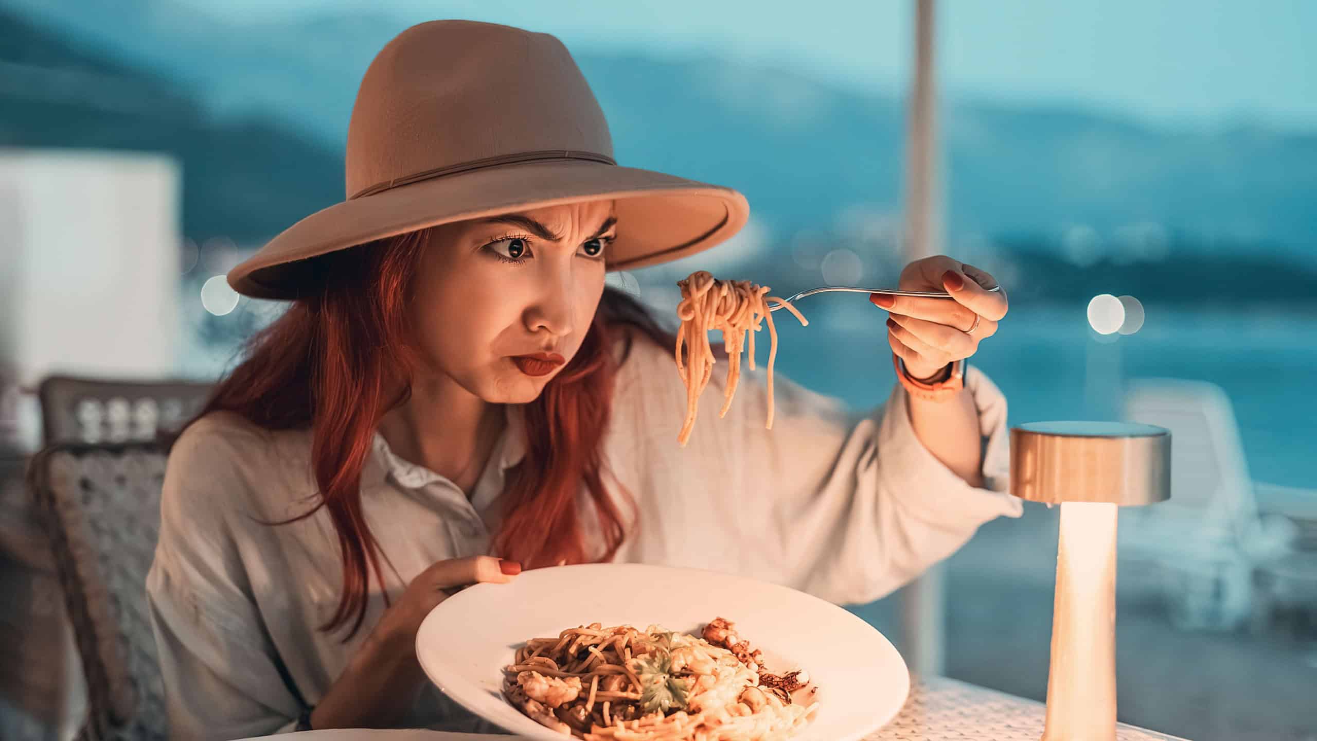 Tourist having dinner in a restaurant with sea view, looking disgusted by her spaghetti