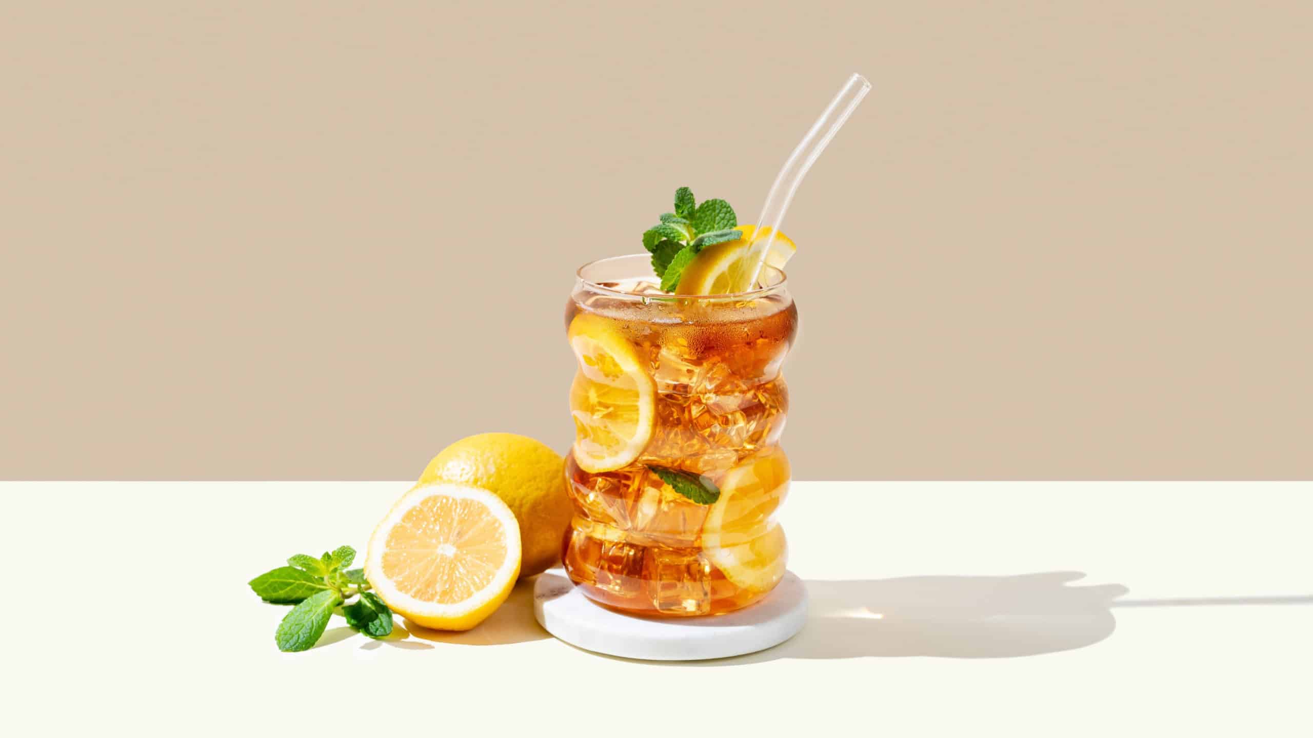 Iced tea with lemon, ice and mint in a glass on beige and white background with shadow and citrus fruits. Trendy concept of refreshing drink or lemonade, menu advertisement mockup. Front view.