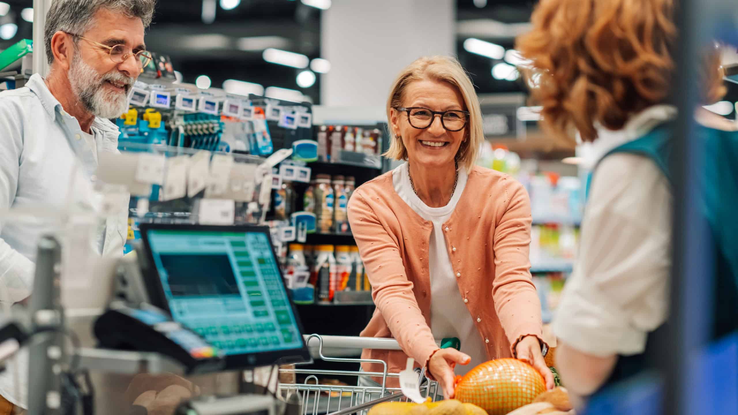 Happy senior woman handing groceries to cashier at checkout counter in supermarket while her husband is waiting beside her
