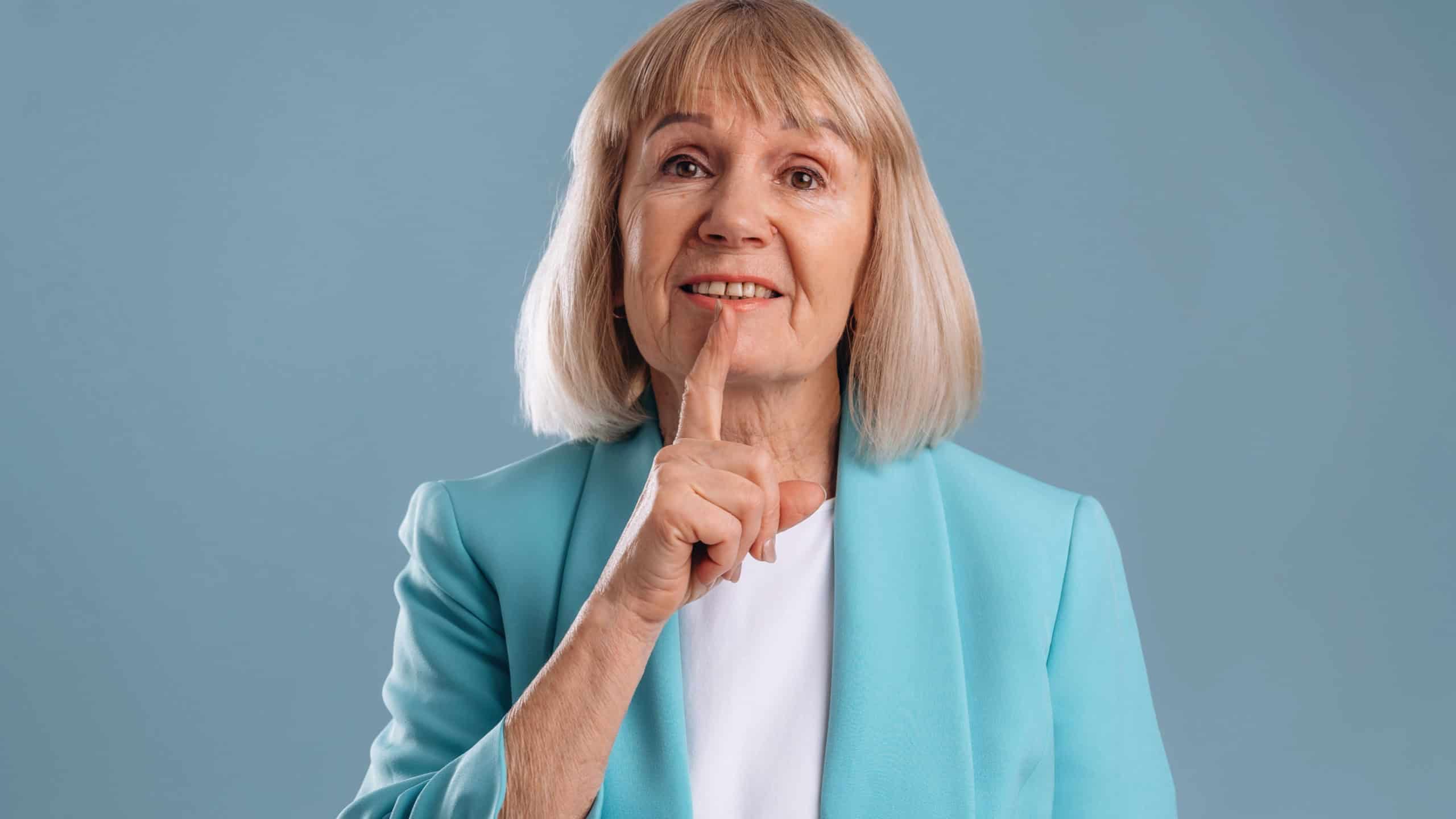 Be silent, finger before the lips. Senior woman is against background.