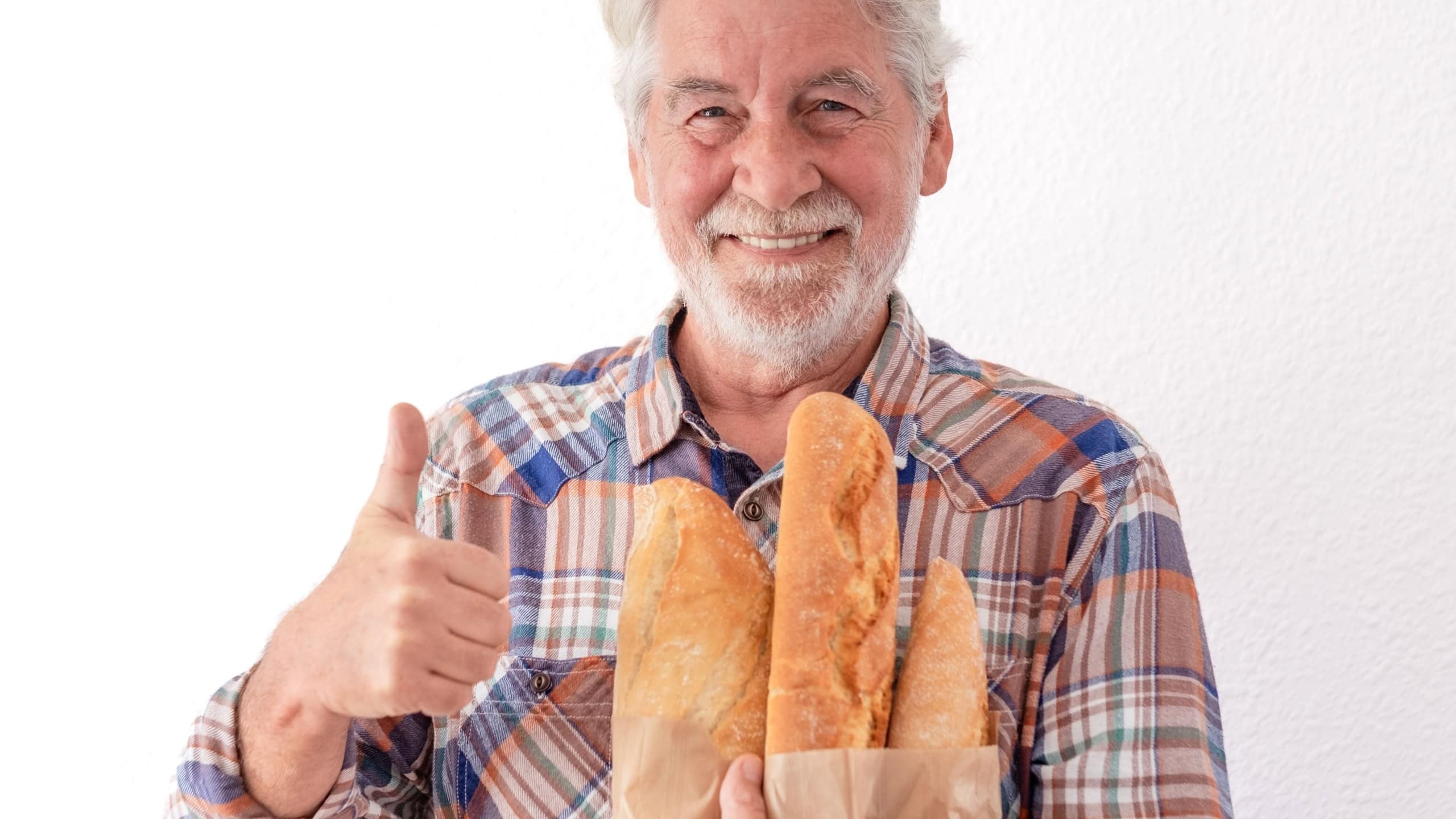 Portrait of Handsome elderly man with white beard holds up a paper bag with just baked bread for the family and looks at camera smiling