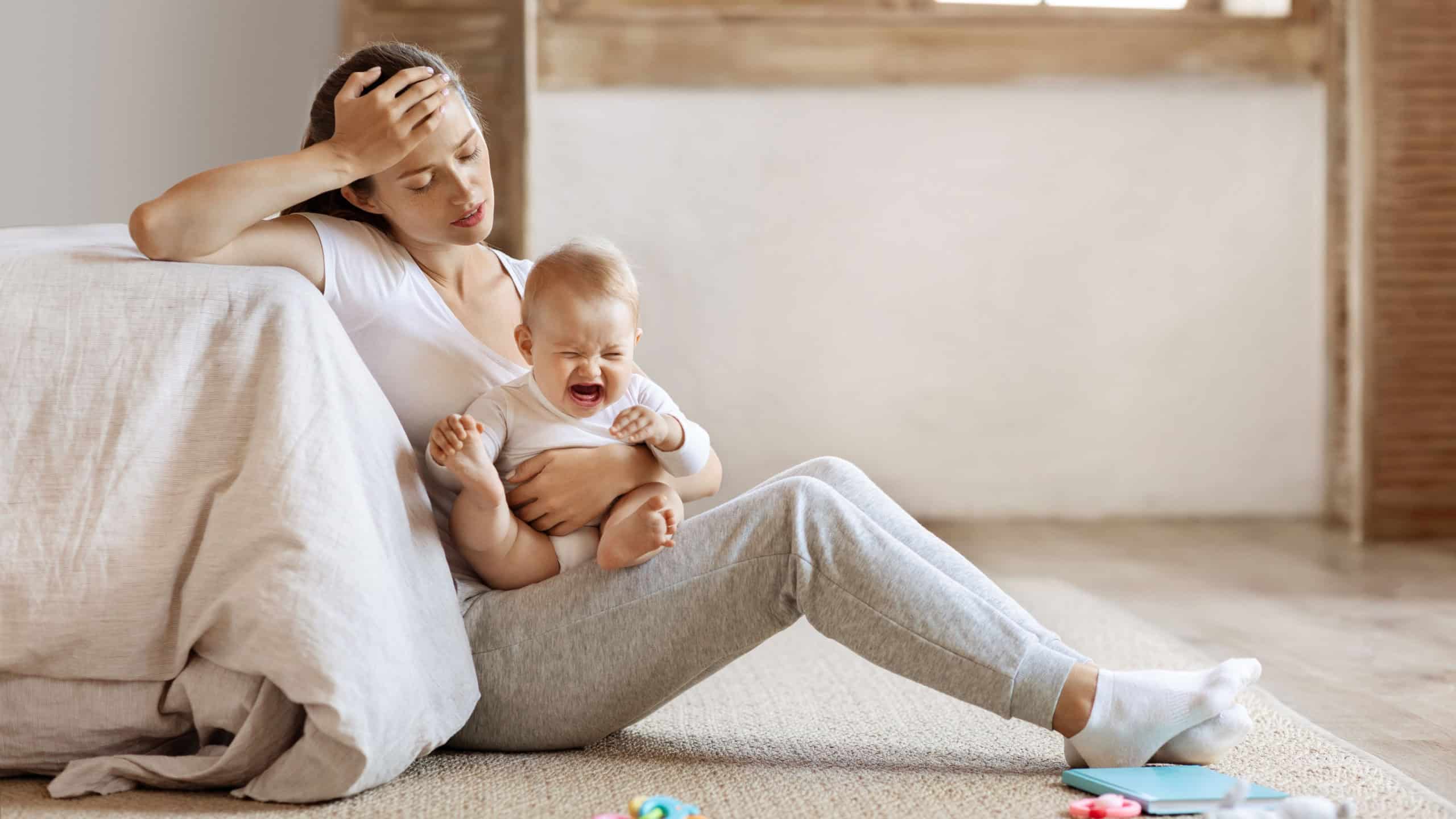 Tired infant baby cries in mother hands, depressed unhappy exhausted mom sitting on floor with crying little child on her lap, bedroom interior, copy space. Postnatal postpartum depression concept