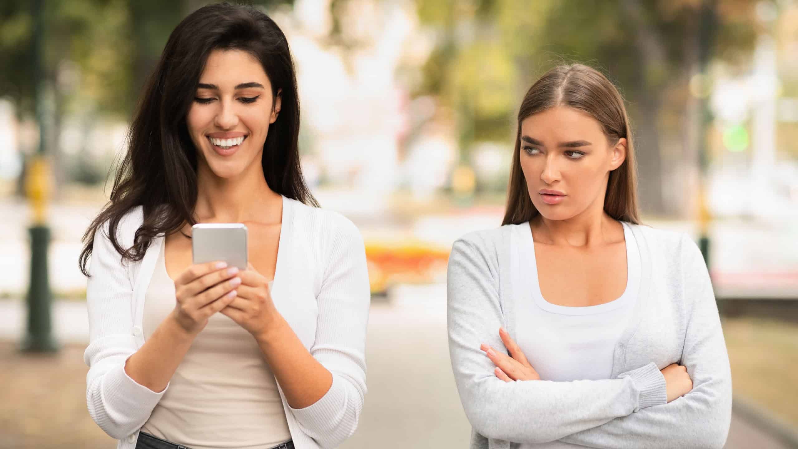 Jealous Girl Looking At Friend's Phone Walking Together Outdoors