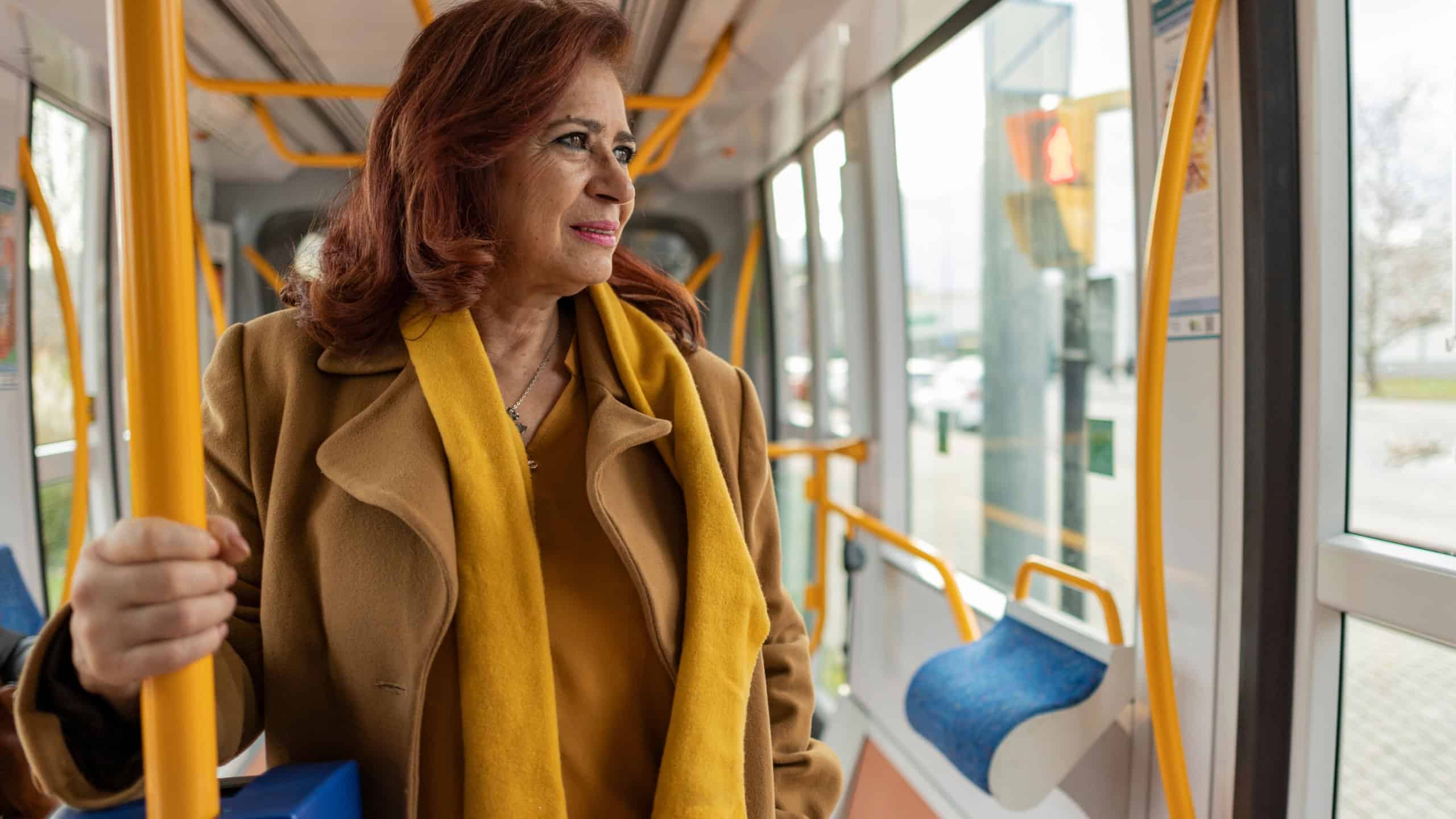 Thoughtful senior woman wearing a yellow scarf and brown coat, standing inside a modern tram or bus, gazing out the window while commuting through the bustling cityscape