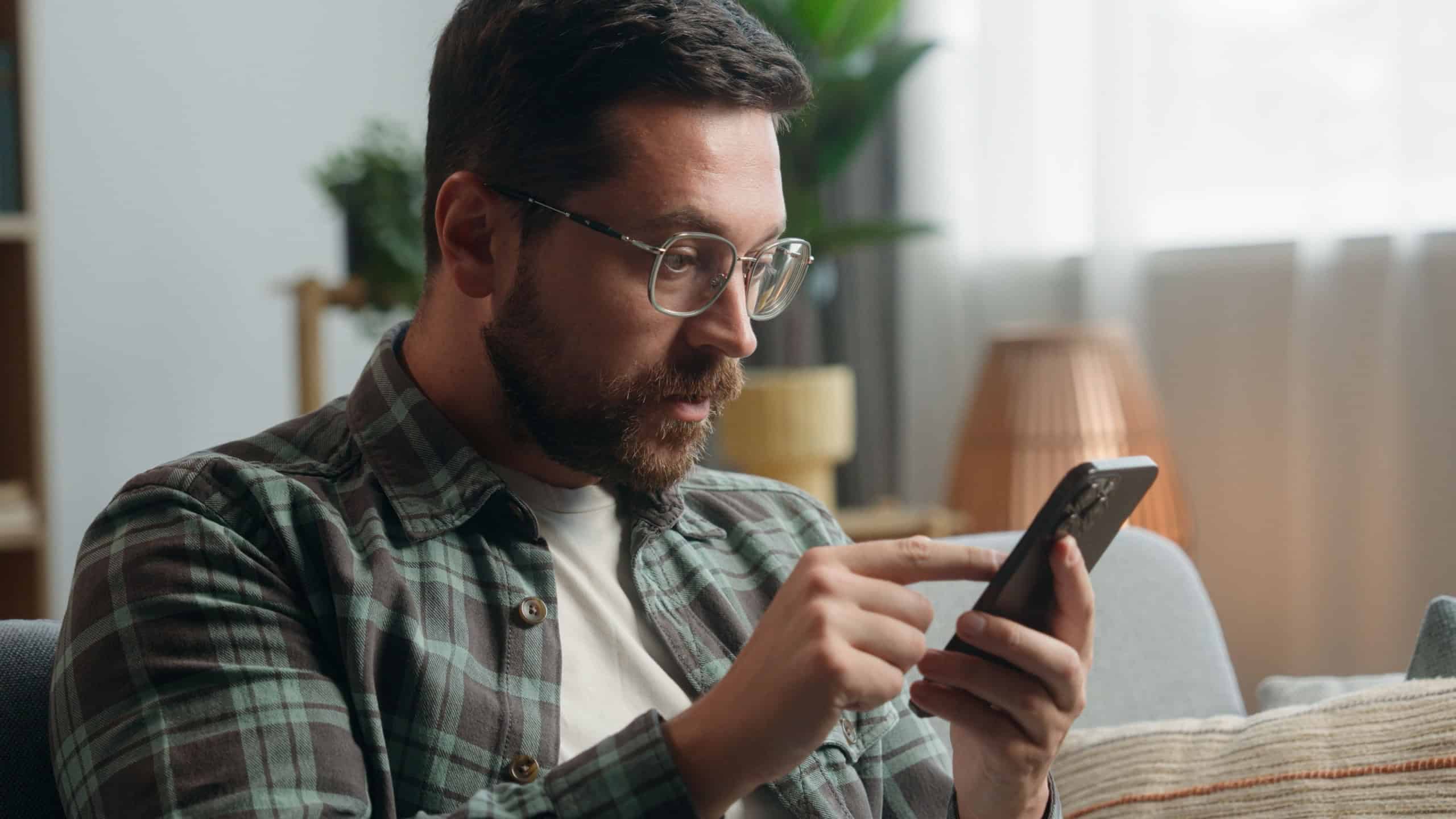 Caucasian man male guy user at home browsing mobile phone app social media smartphone shock bad news amazed trouble problem confused puzzled wonder online internet web at home couch sofa apartment