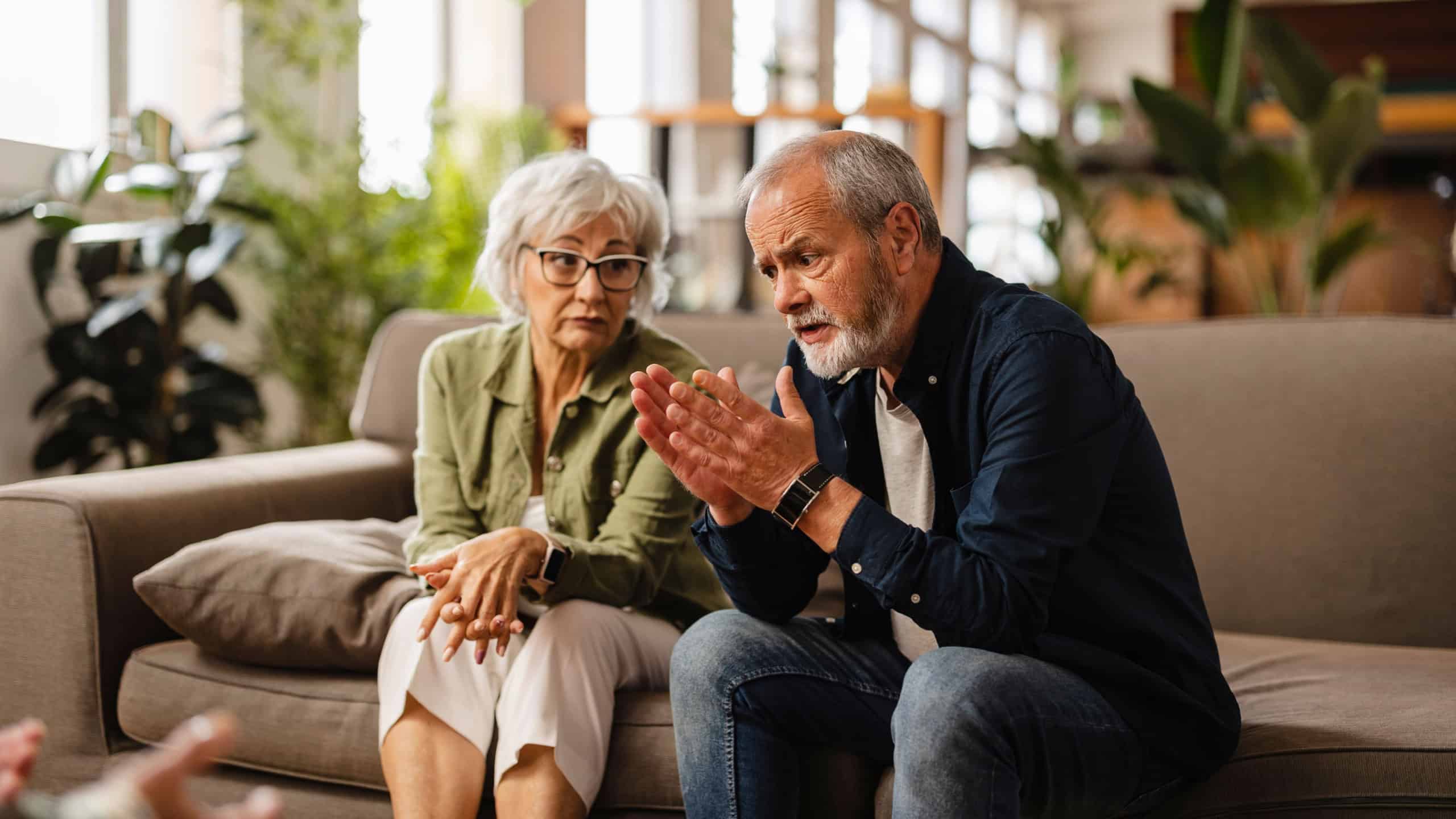 Senior couple sitting on a sofa, discussing their problems with a psychologist during a therapy session at home, sharing concerns and seeking guidance for their relationship issues