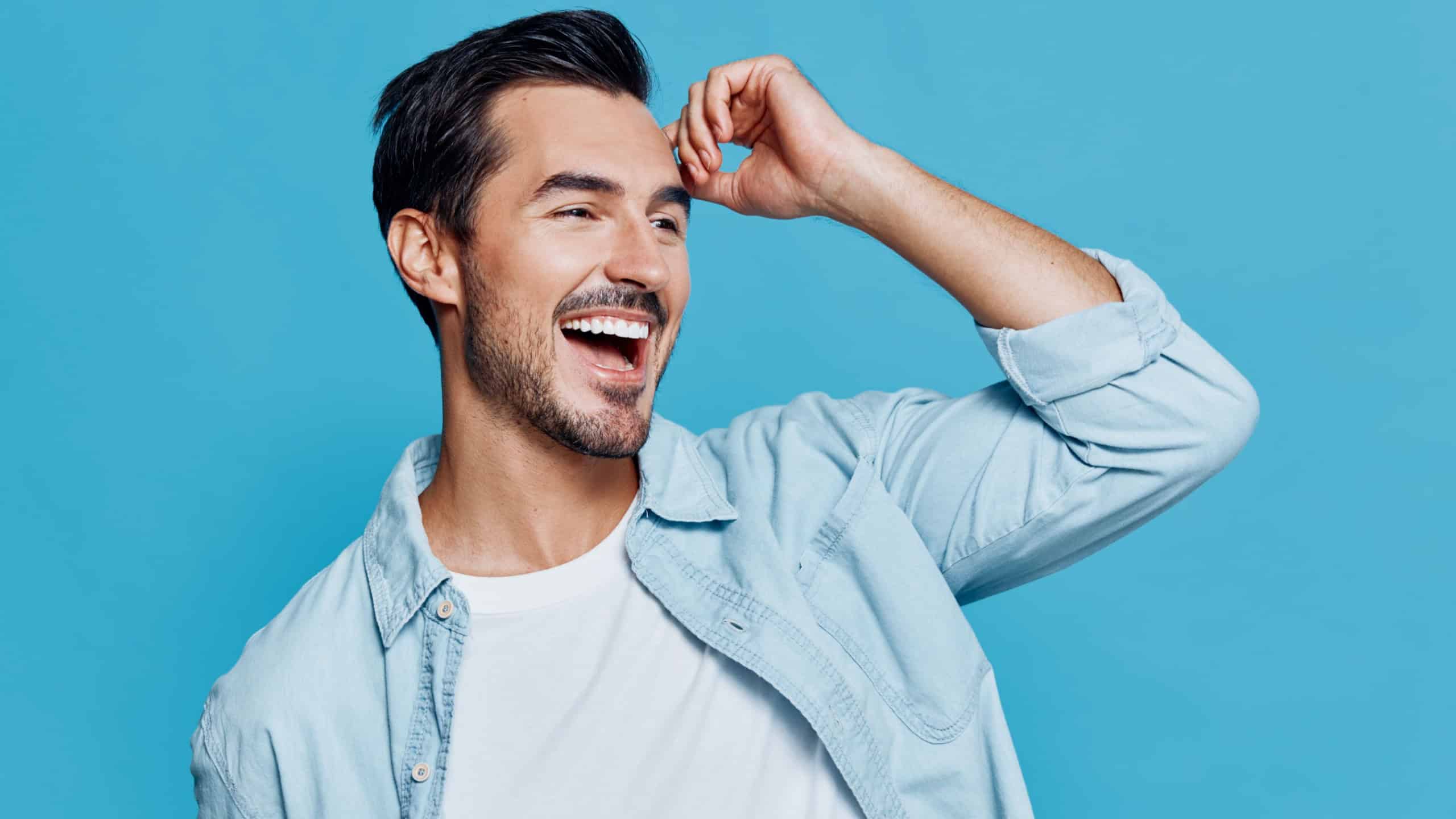 Handsome smiling man with stylish hair wearing a light blue denim shirt, posing joyfully against a vibrant blue background. Cheerful expression concept.