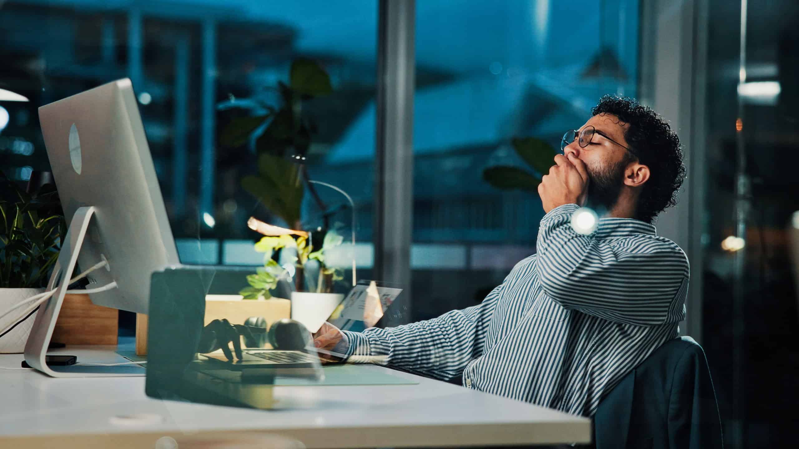 Tired, businessman and night with yawn on computer for project deadline or exhausted in office. Man, employee or workaholic working late with technology for fatigue, insomnia or overtime in workplace