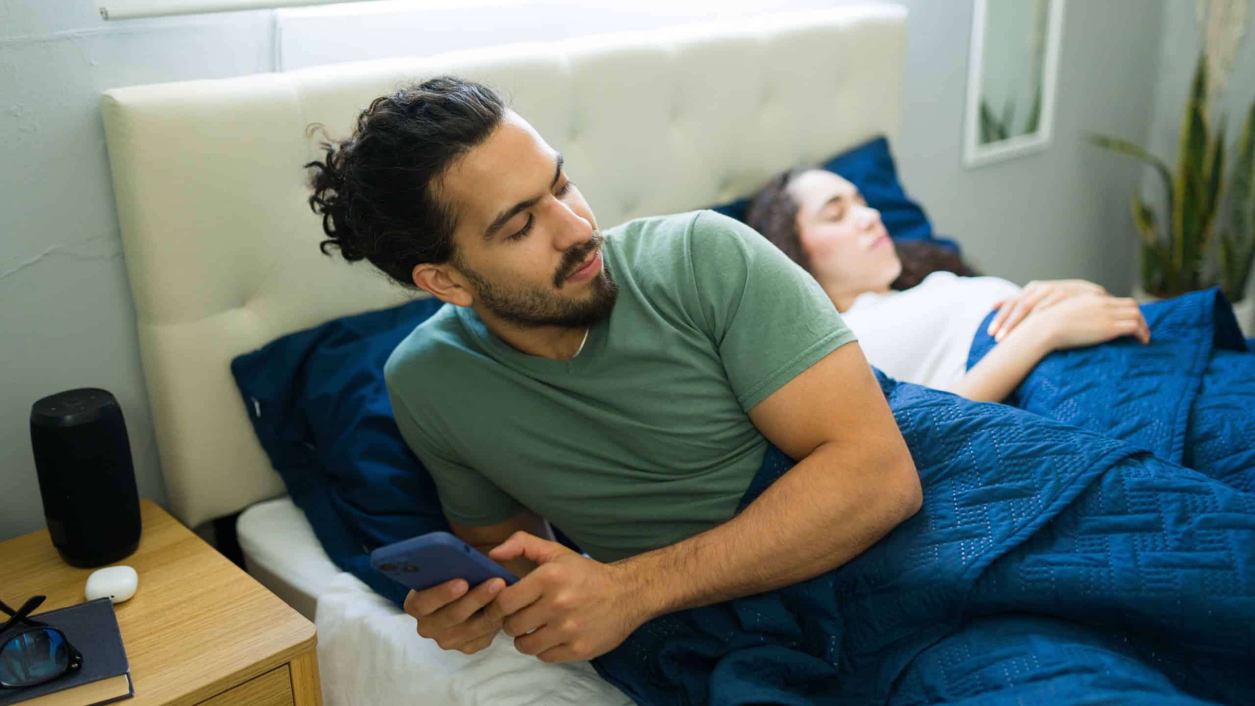 Young man using smartphone and hiding activity from his partner sleeping next to him in bed at home