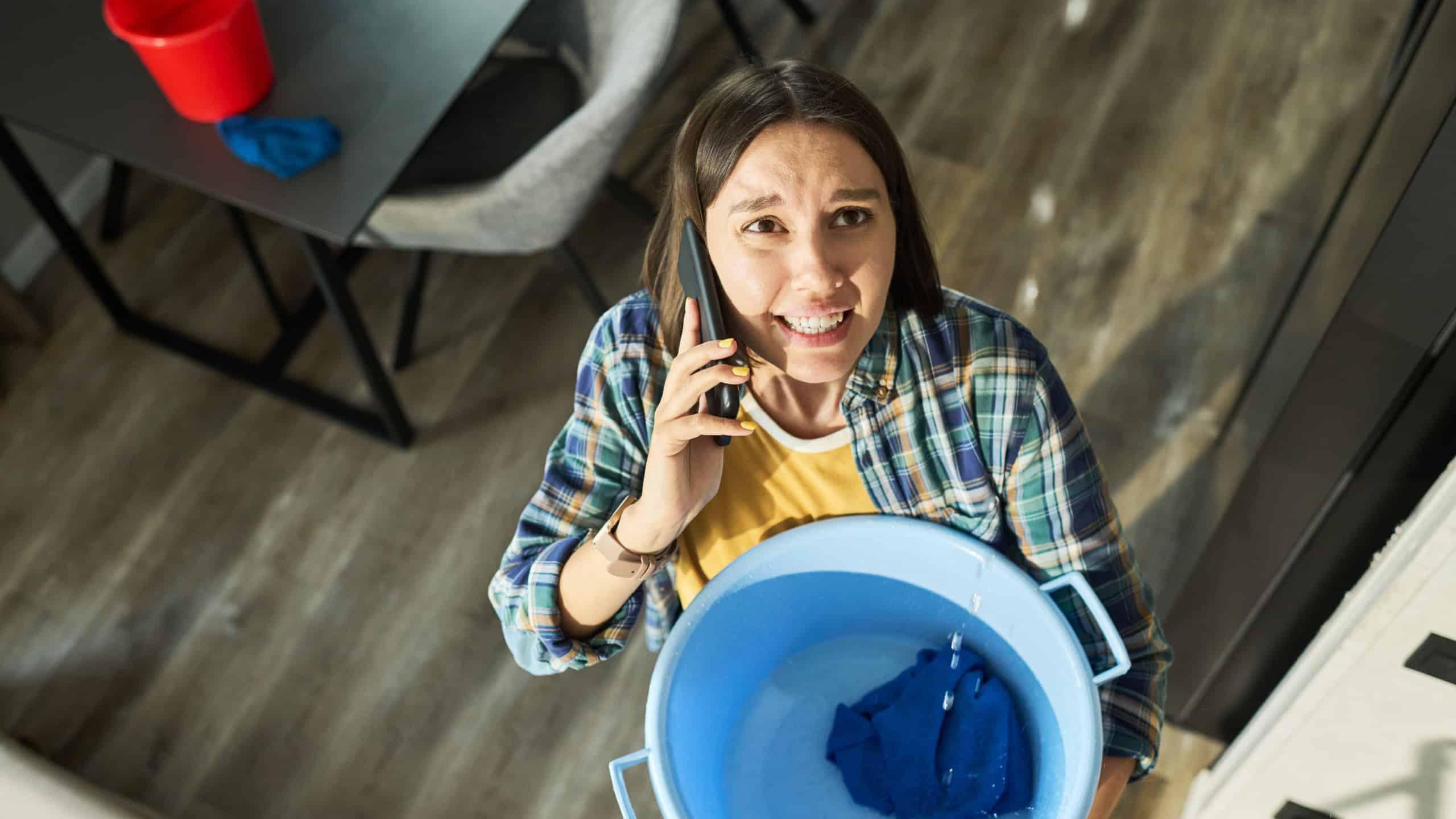 Caucasian young adult woman standing indoors holding blue plastic basin with water and cloth while talking on smartphone looking up with worried expression