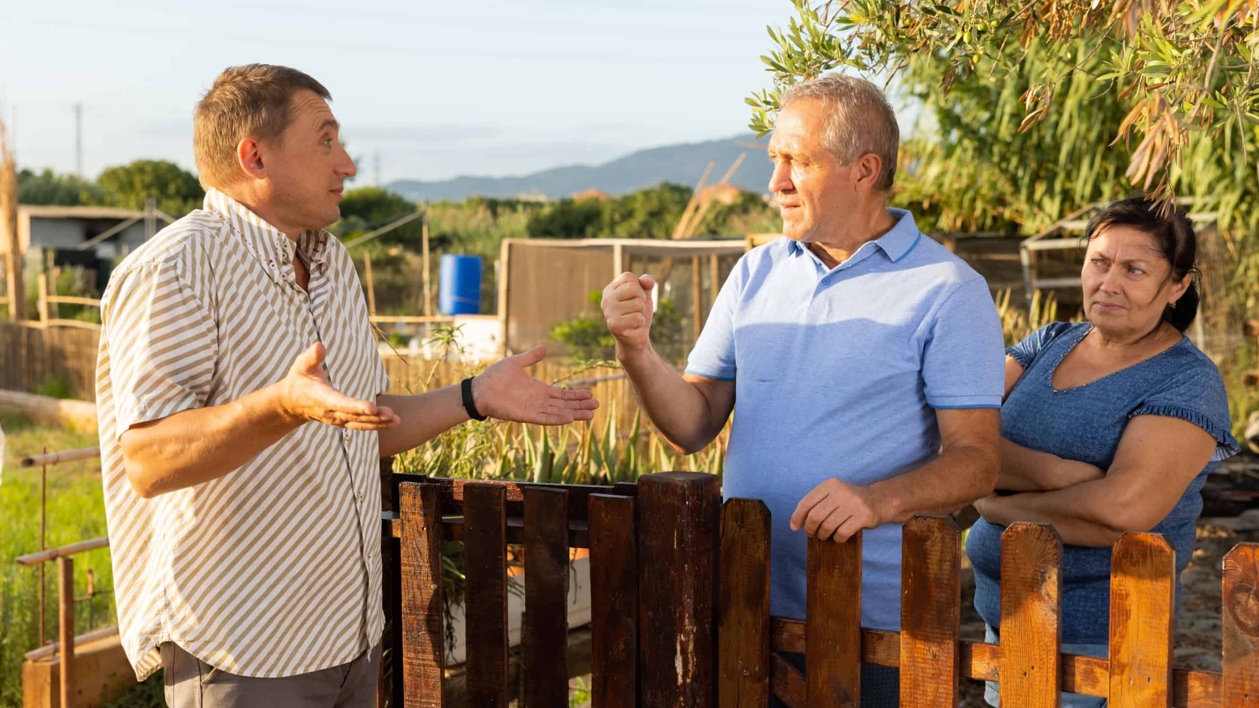 Portrait of man standing at fence and having quarrel with mature couple neighbours at backyard garden. Mature man frightening neighbor with fist gesture.