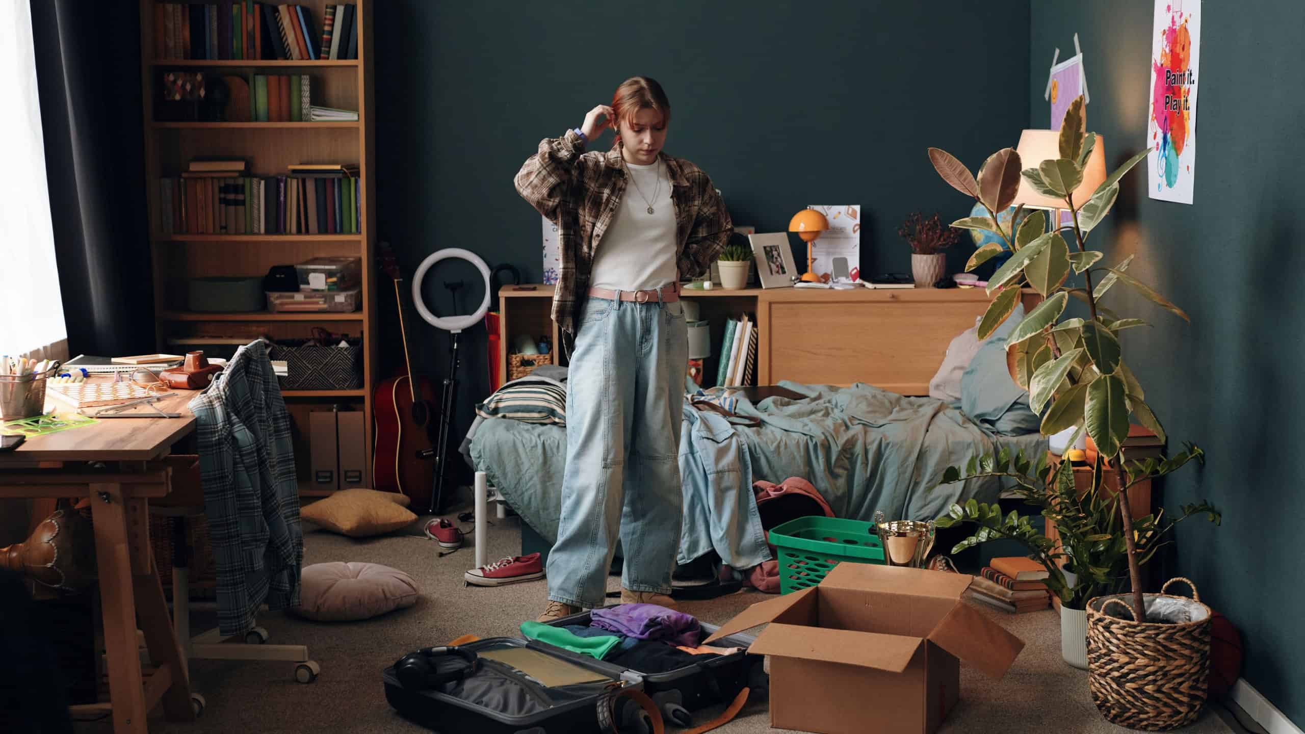 Caucasian teenage girl standing in messy bedroom packing suitcase and cardboard box, organizing clothes and personal items, surrounded by books and plants, appearing thoughtful