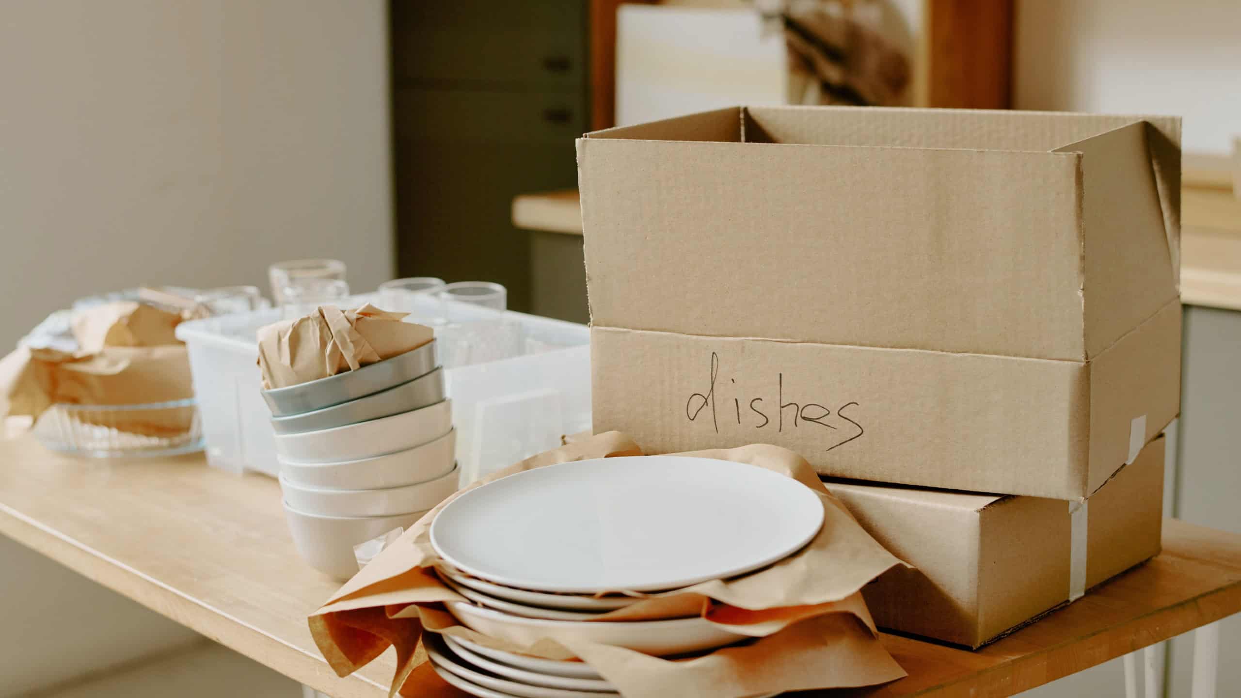 Stacked cardboard boxes labeled dishes and neatly arranged plates, bowls, and glasses on wooden table, showing unpacking process during young couple moving and doing renovations