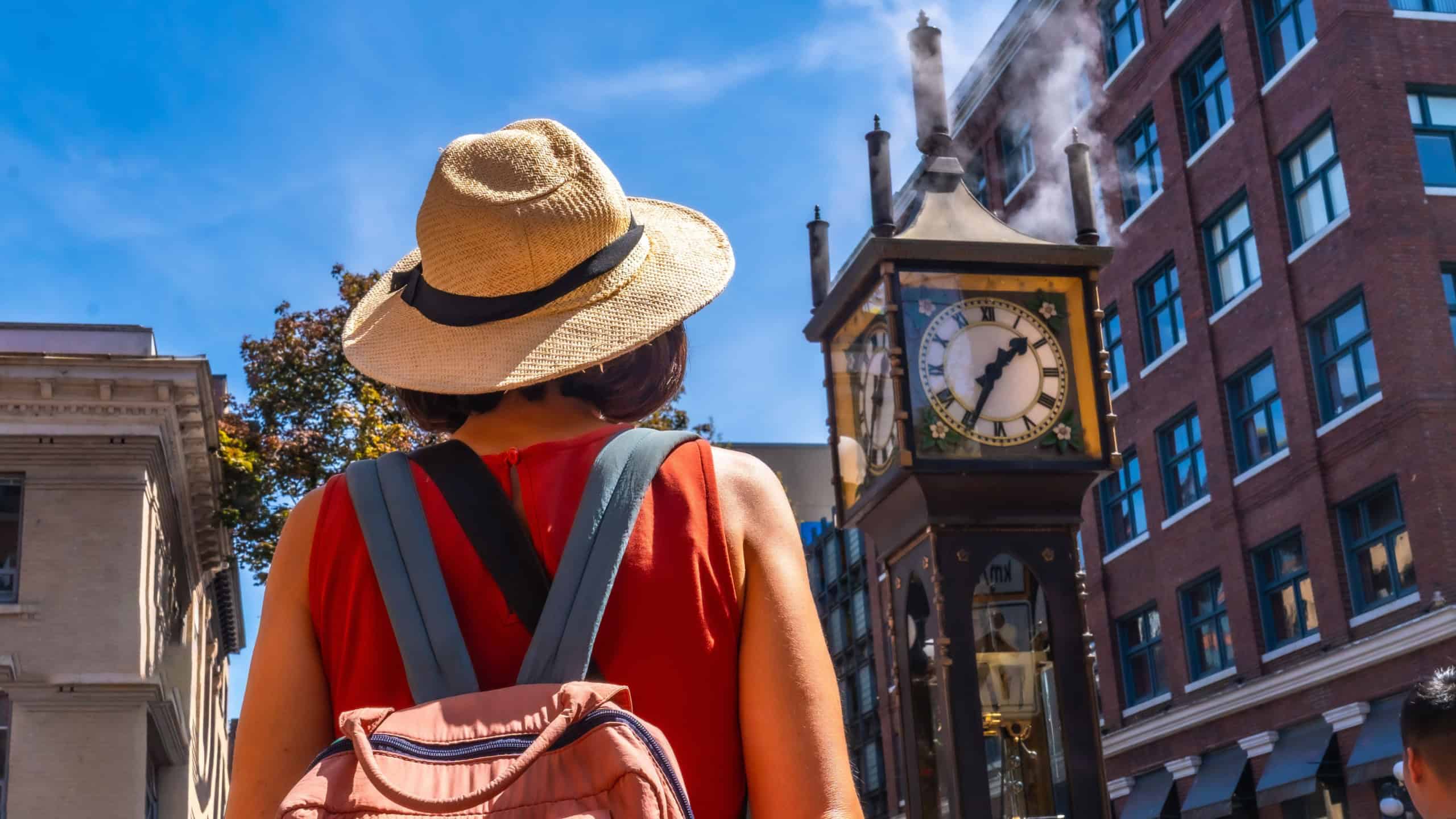 Female tourist wearing straw hat and backpack looking at the famous steam clock in gastown historic district, vancouver, british columbia, canada, in a sunny summer day