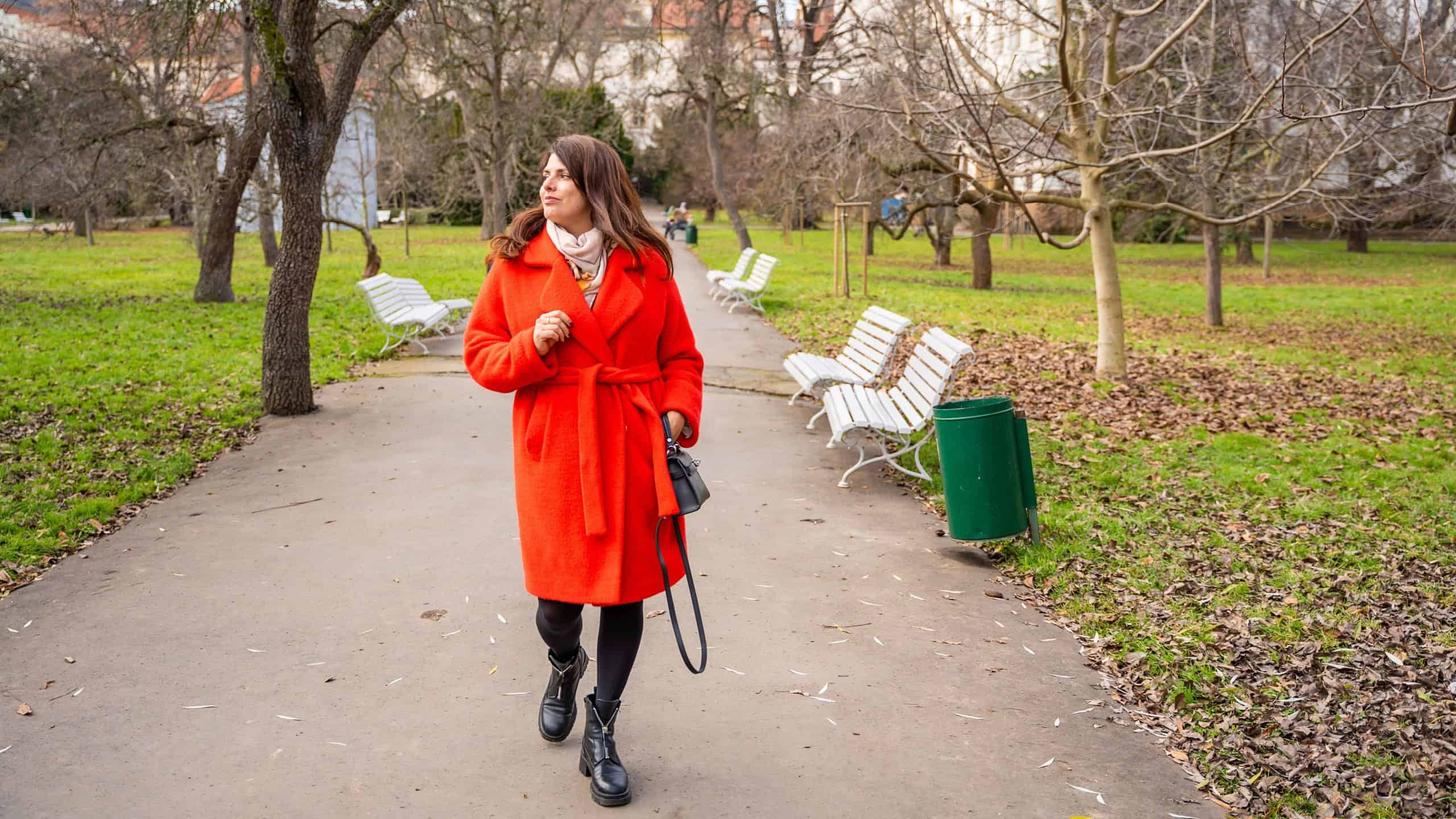 Woman walking in park in Prague during winter without snow wearing red coat. Calm urban walking, everyday lifestyle and seasonal city routine in real life European environment.