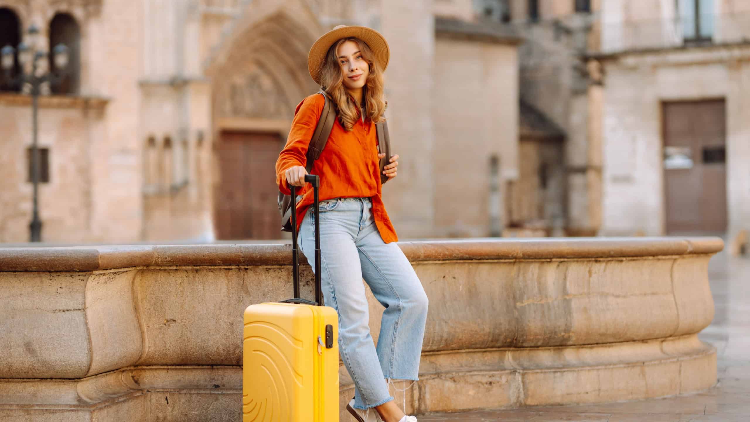 Female tourist wearing hat and carrying yellow suitcase strolls through sunny streets of city. Happy woman enjoys adventure with small suitcase and explores new sights. Tourism concept, weekend.