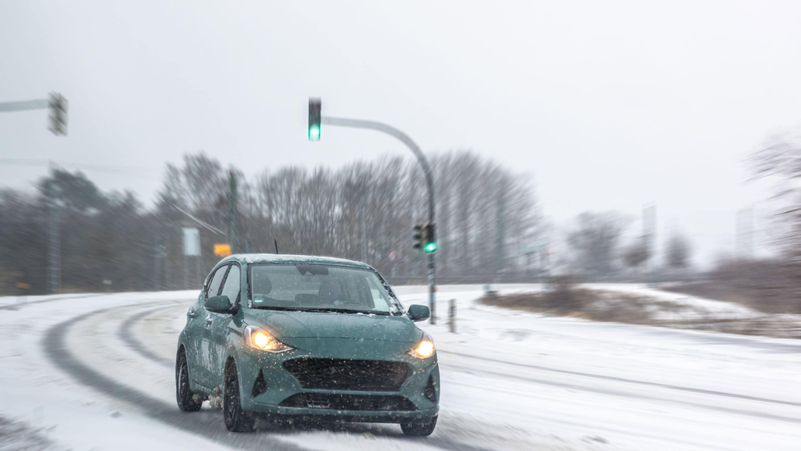 A small car with headlights turns on a snow covered road with a green light glowing in cold haze. The frame highlights urban mobility, visibility, and cautious driving.