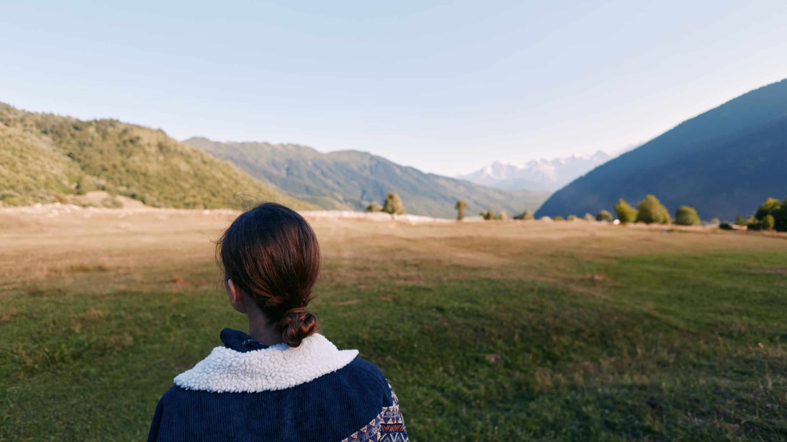 person woman mountains valley meadow nature back landscape in this photo: a woman seen from behind stands in a wide grassy meadow looking toward distant mountain valley under clear sky