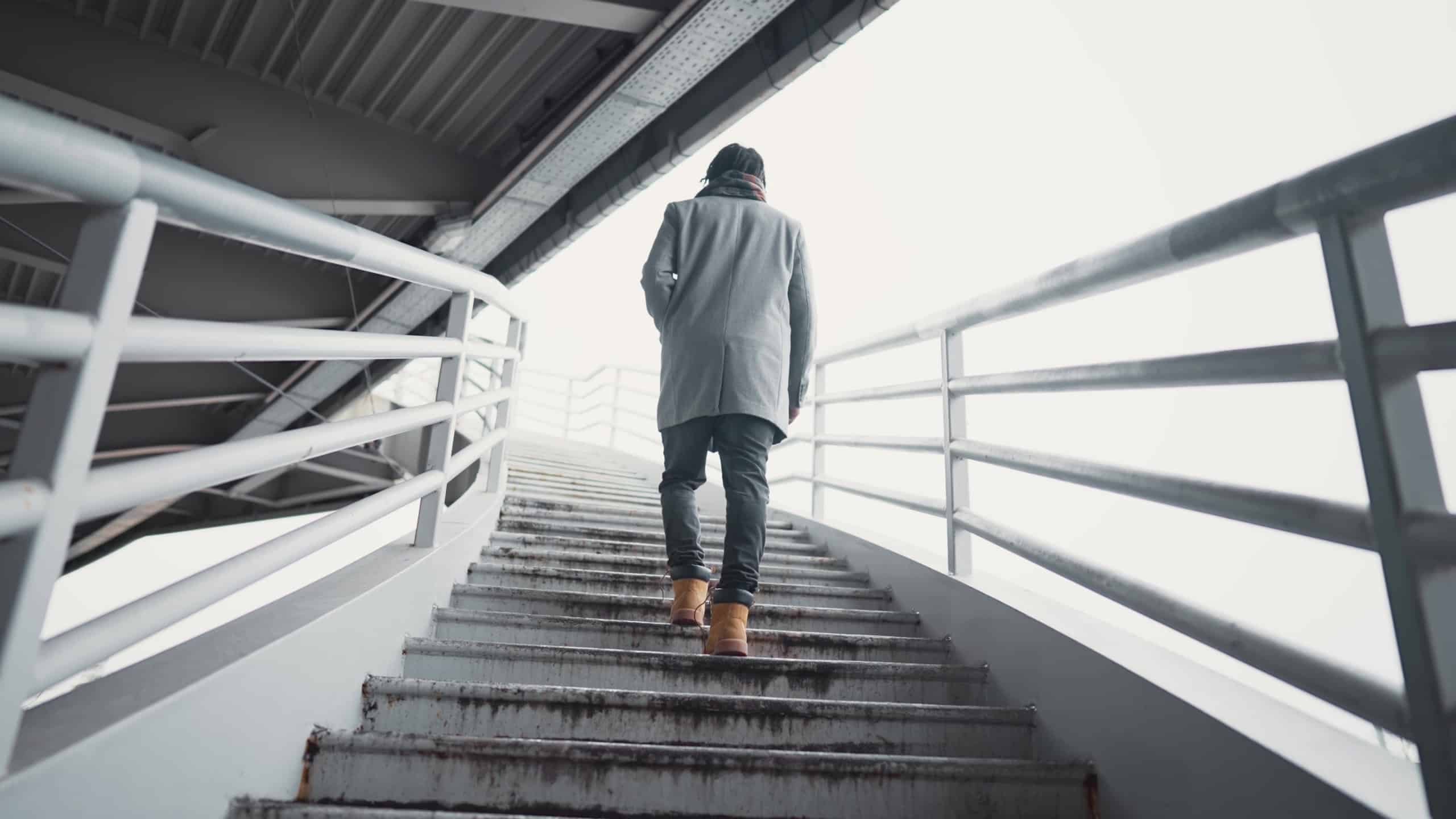 Lonely male person walking in urbanscape, back view of man moving up on stairs. Slow motion shot, african american guy strolling over metal ladder, back view, warm clothes for autumn season in city